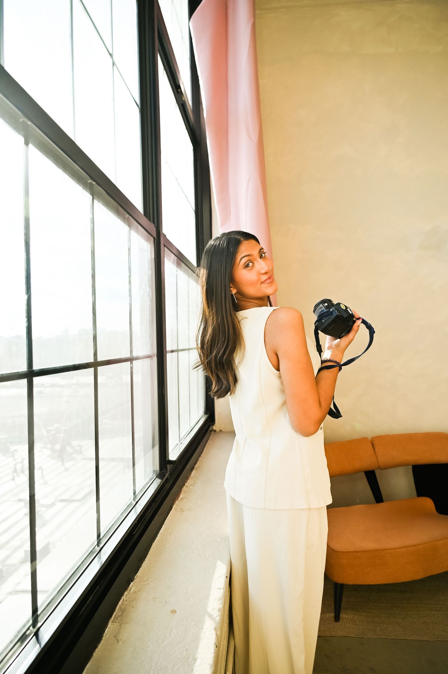 Woman in white dress poses by large windows in elegant indoor photoshoot.