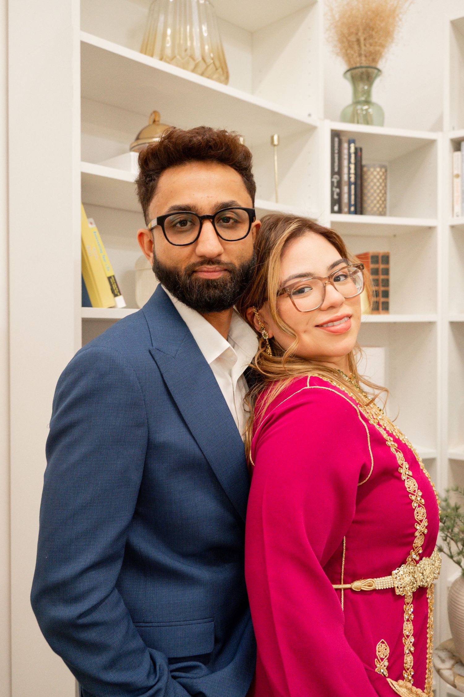 A couple poses together in front of white built-in shelves with decorative items.