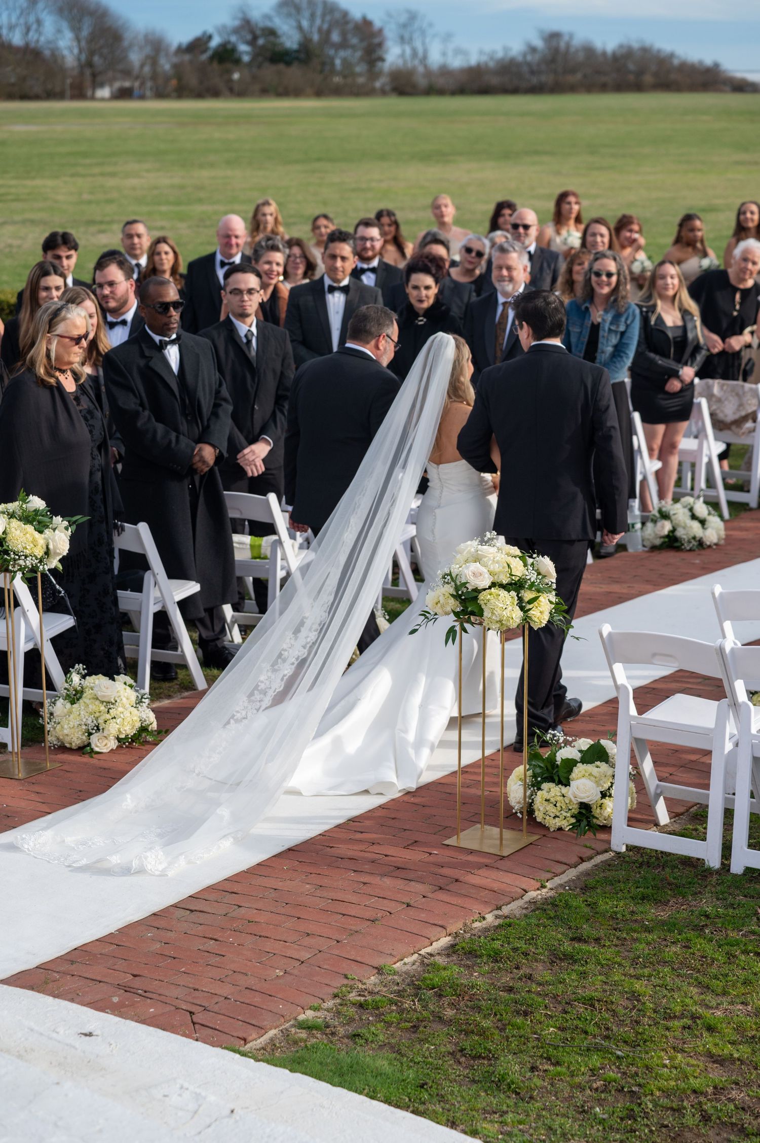 A white aisle runner unfurls along a brick pathway at an outdoor wedding ceremony with guests seated on white chairs.