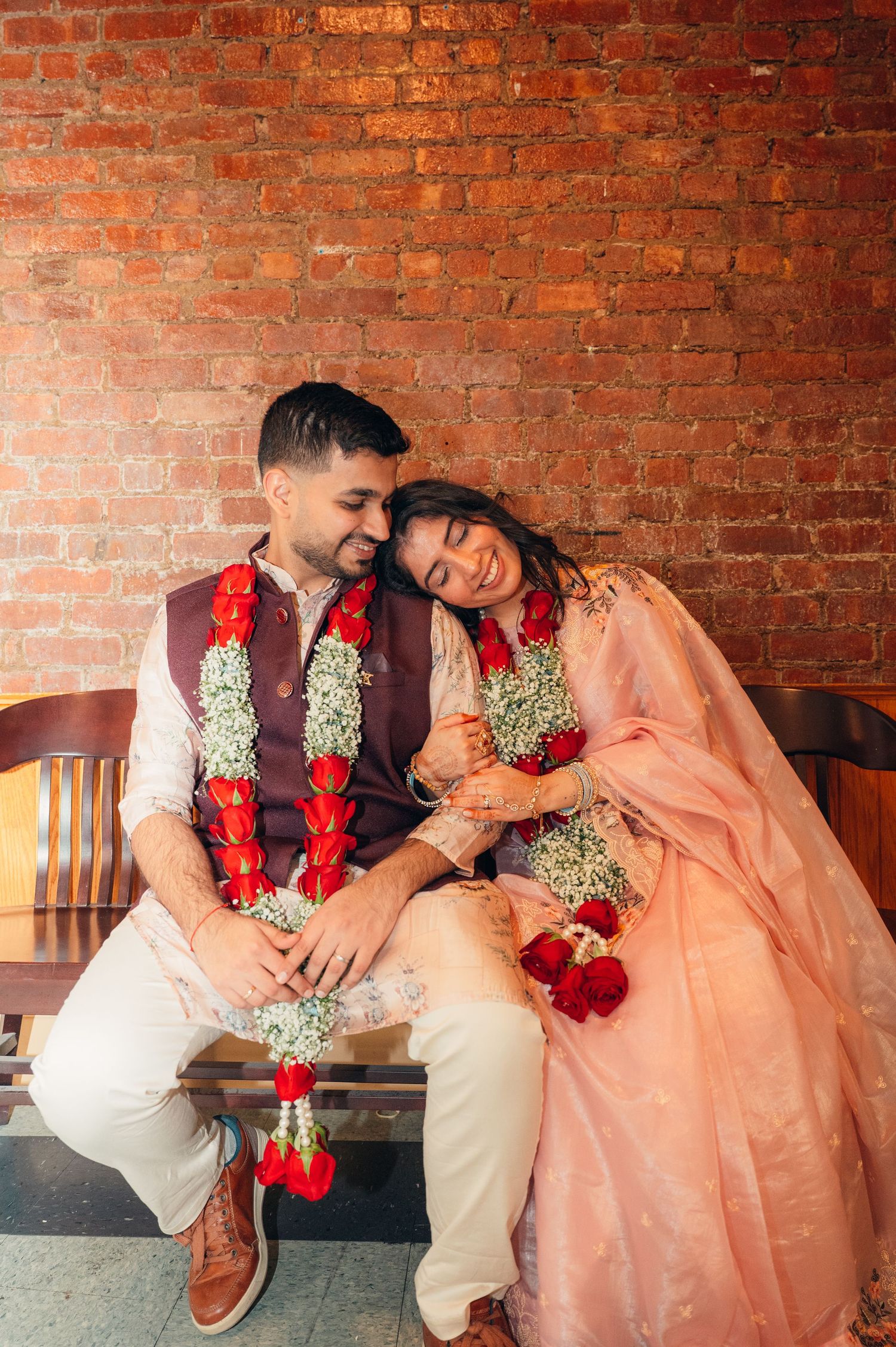 A romantic couple in traditional Indian wedding attire sits together against a rustic brick wall background.