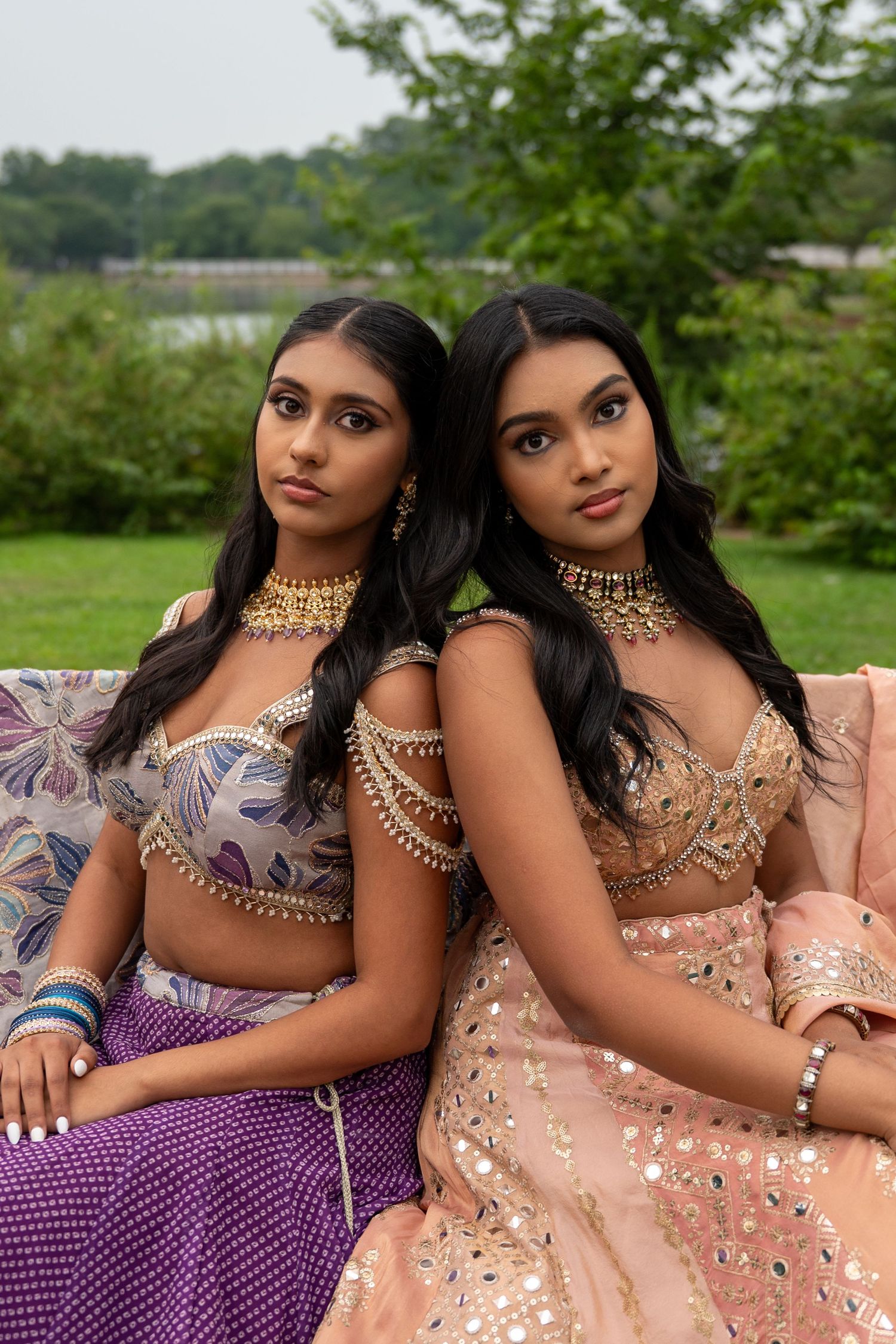 Two women in ornate Indian formal wear pose together on outdoor sofa.