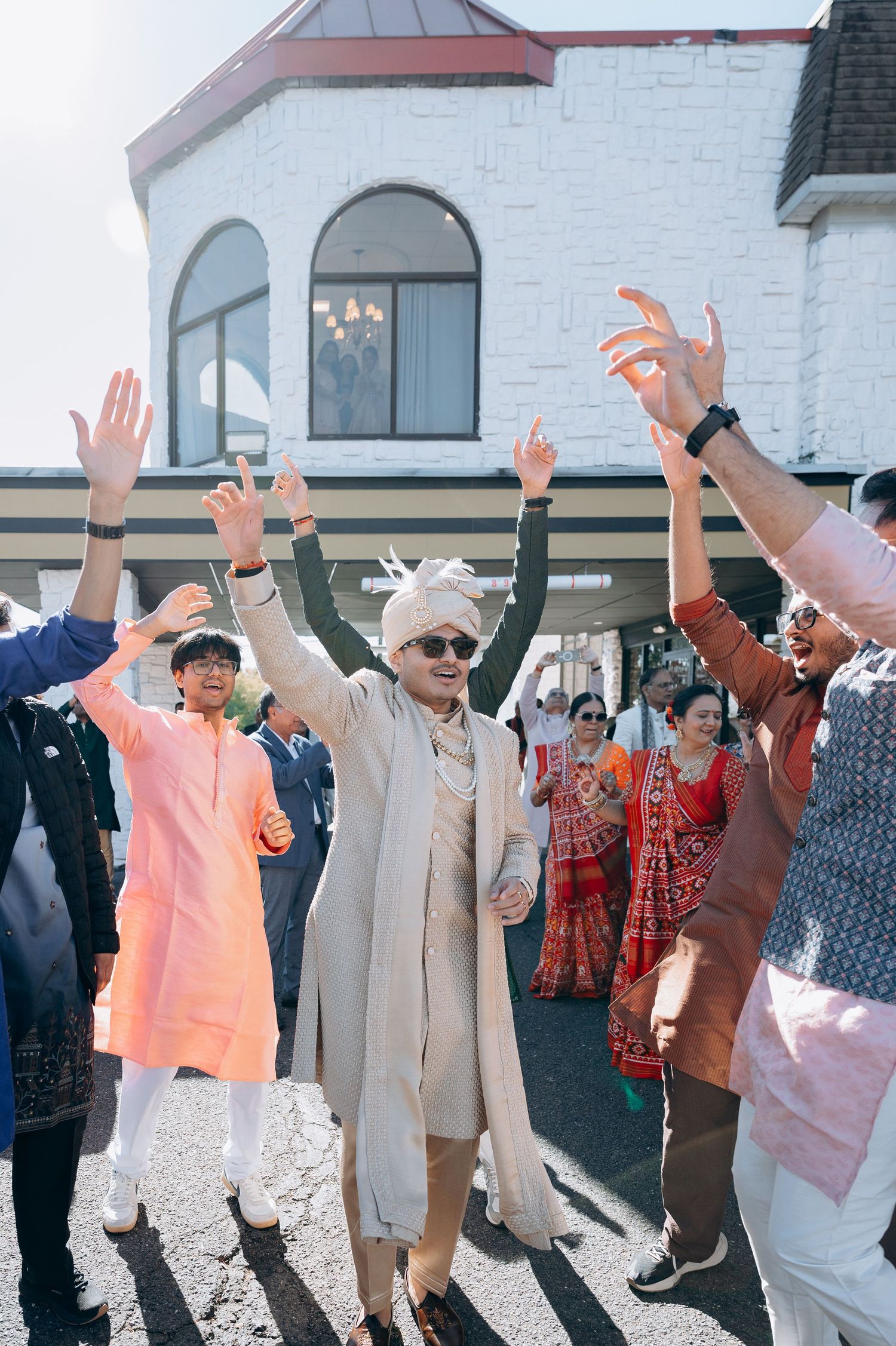 Wedding guests in traditional Indian attire raising their hands in celebration outside.