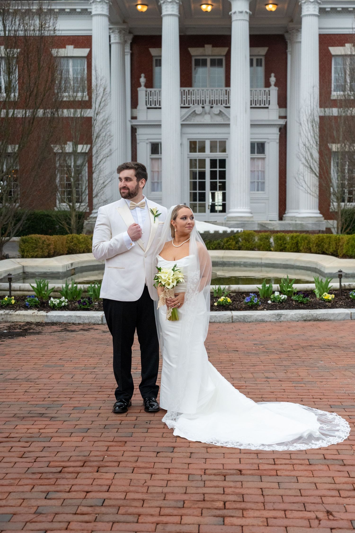 Wedding couple posing on red brick courtyard in front of white columned building.