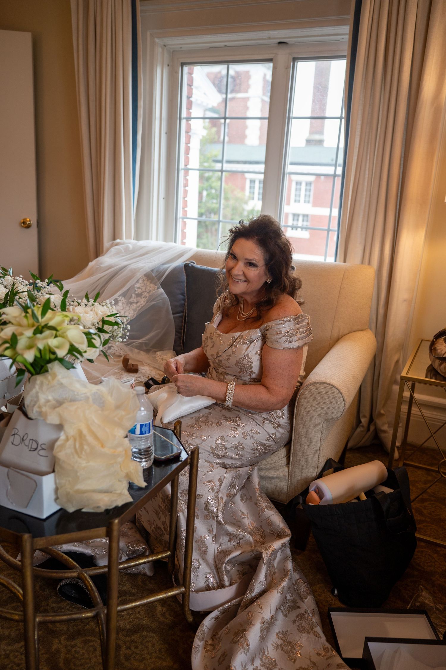 A bride in a lace wedding dress sits in an elegant room with white floral arrangements and large windows.