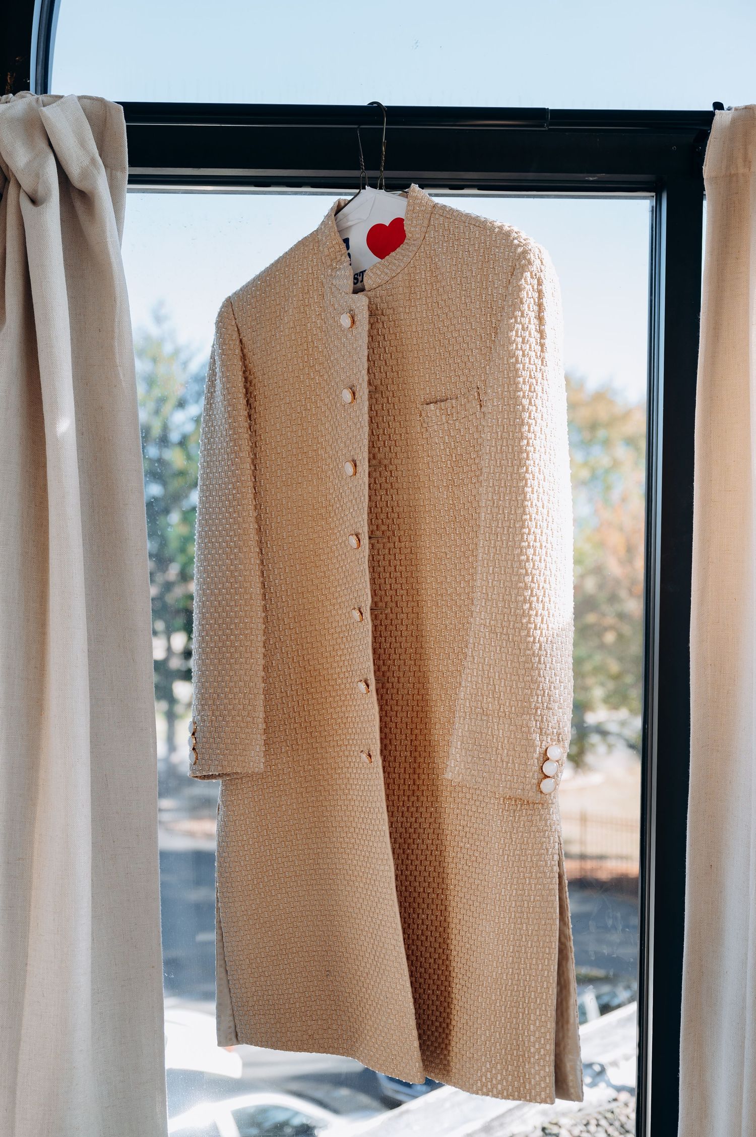 Beige formal suits hanging on hangers by a window with natural lighting.