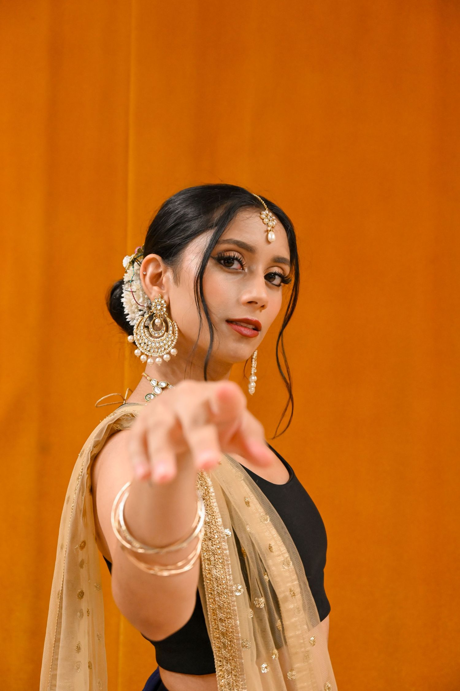 A woman in traditional South Asian attire poses against an orange backdrop wearing ornate jewelry and a dupatta.