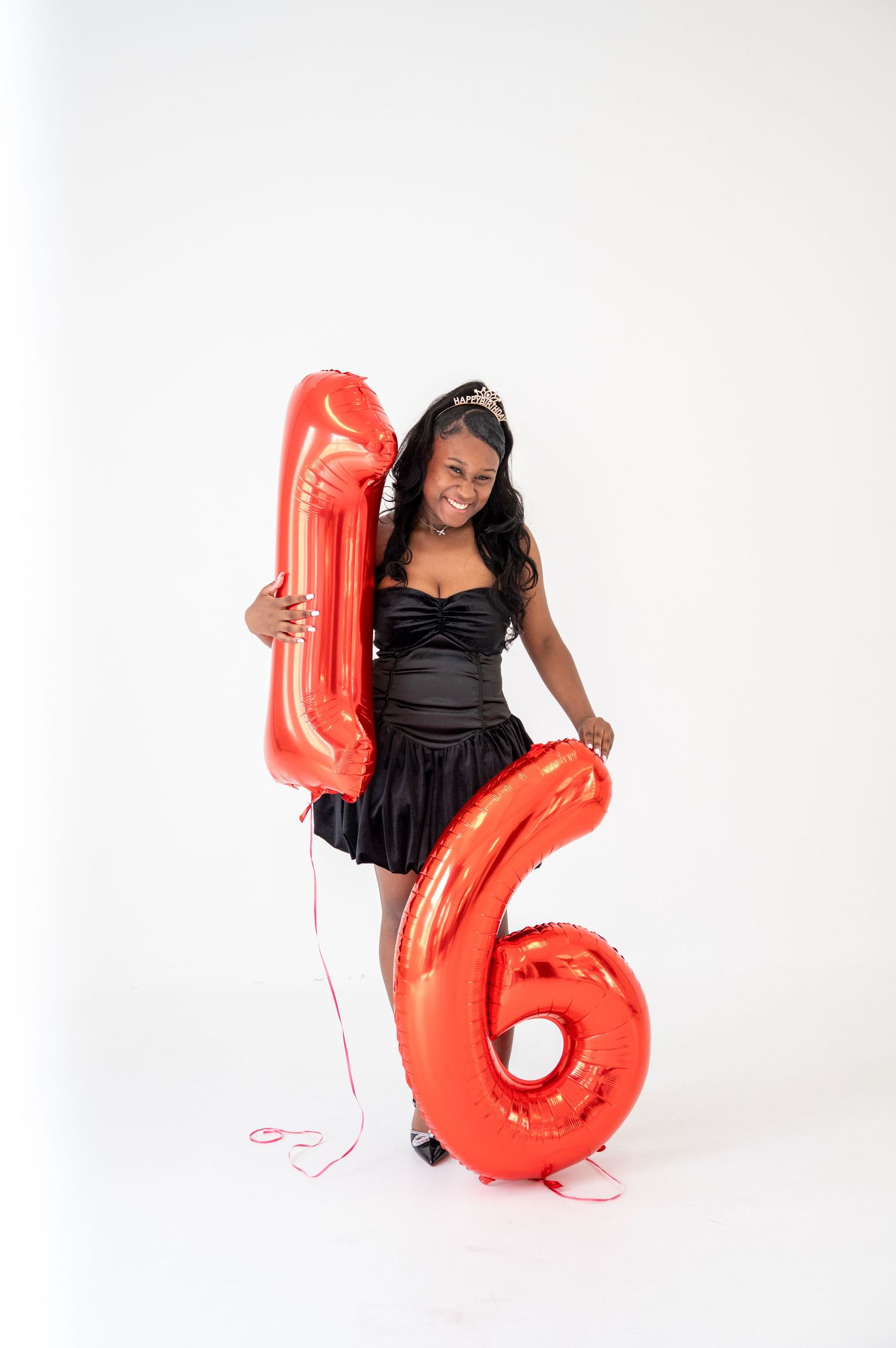 Person in black dress poses with large red number 16 balloon on white background.