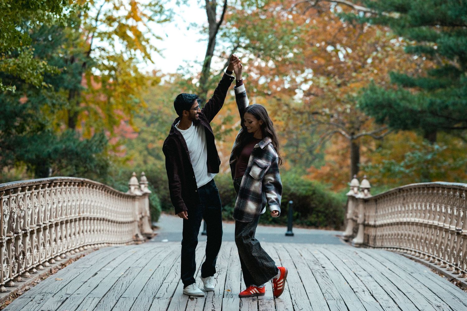Couple walking across a scenic wooden bridge surrounded by autumn foliage in a park setting.