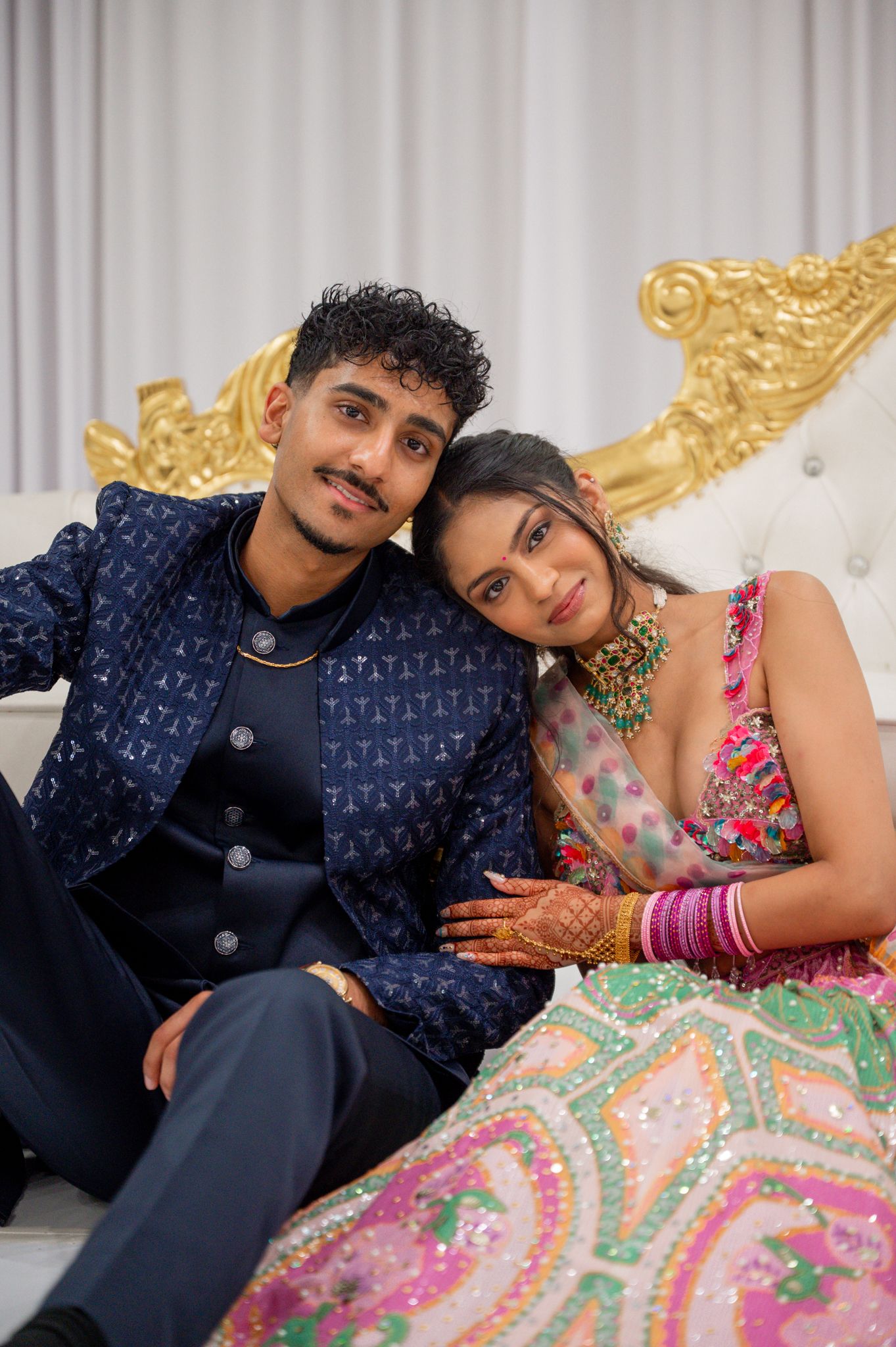 Indian couple in traditional wedding attire, man in navy sherwani and woman in colorful lehenga with mehndi, posing on ornate white sofa.