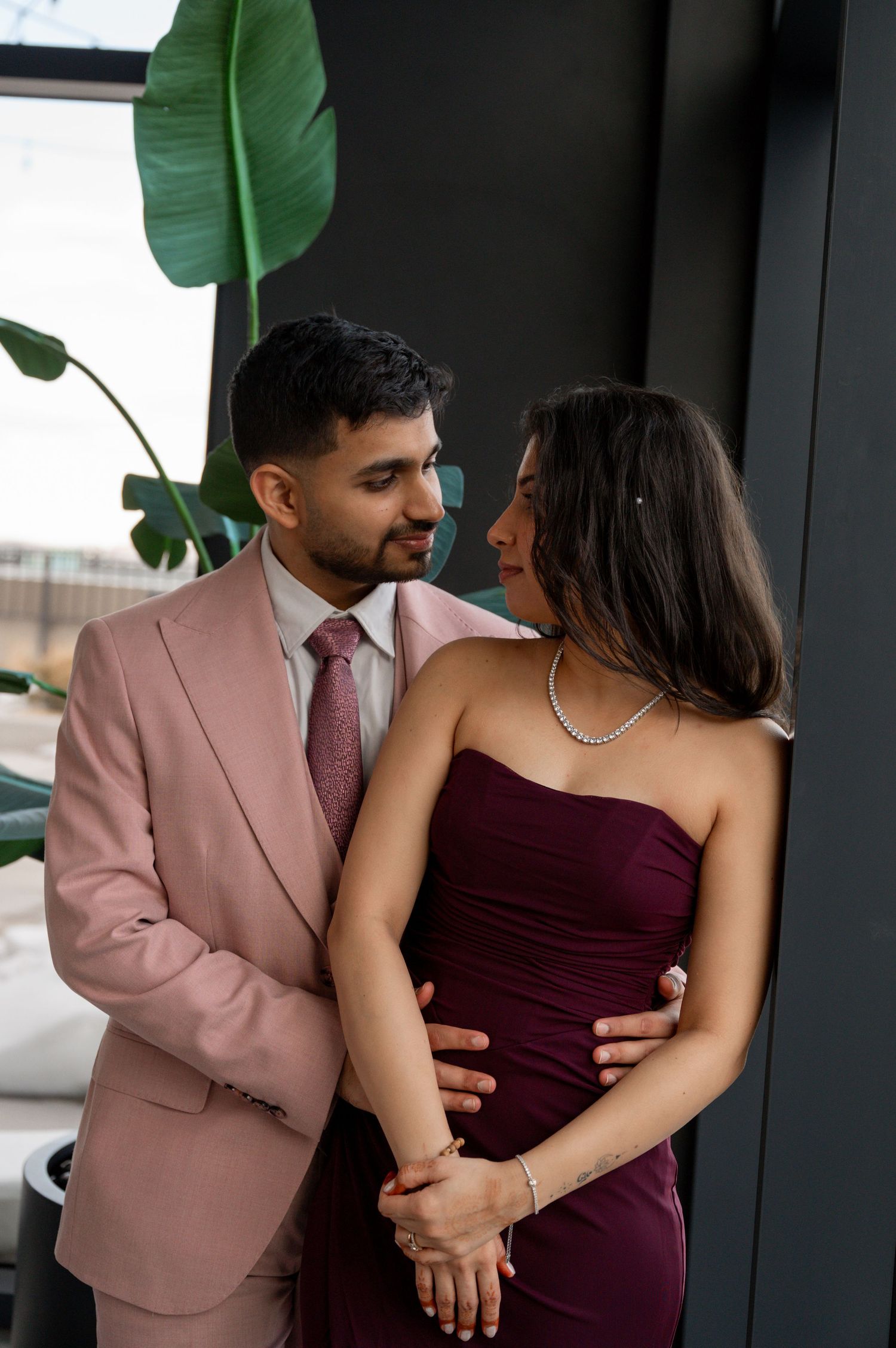 Couple in formal attire embrace near indoor plant, with woman in burgundy dress and man wearing pink suit.