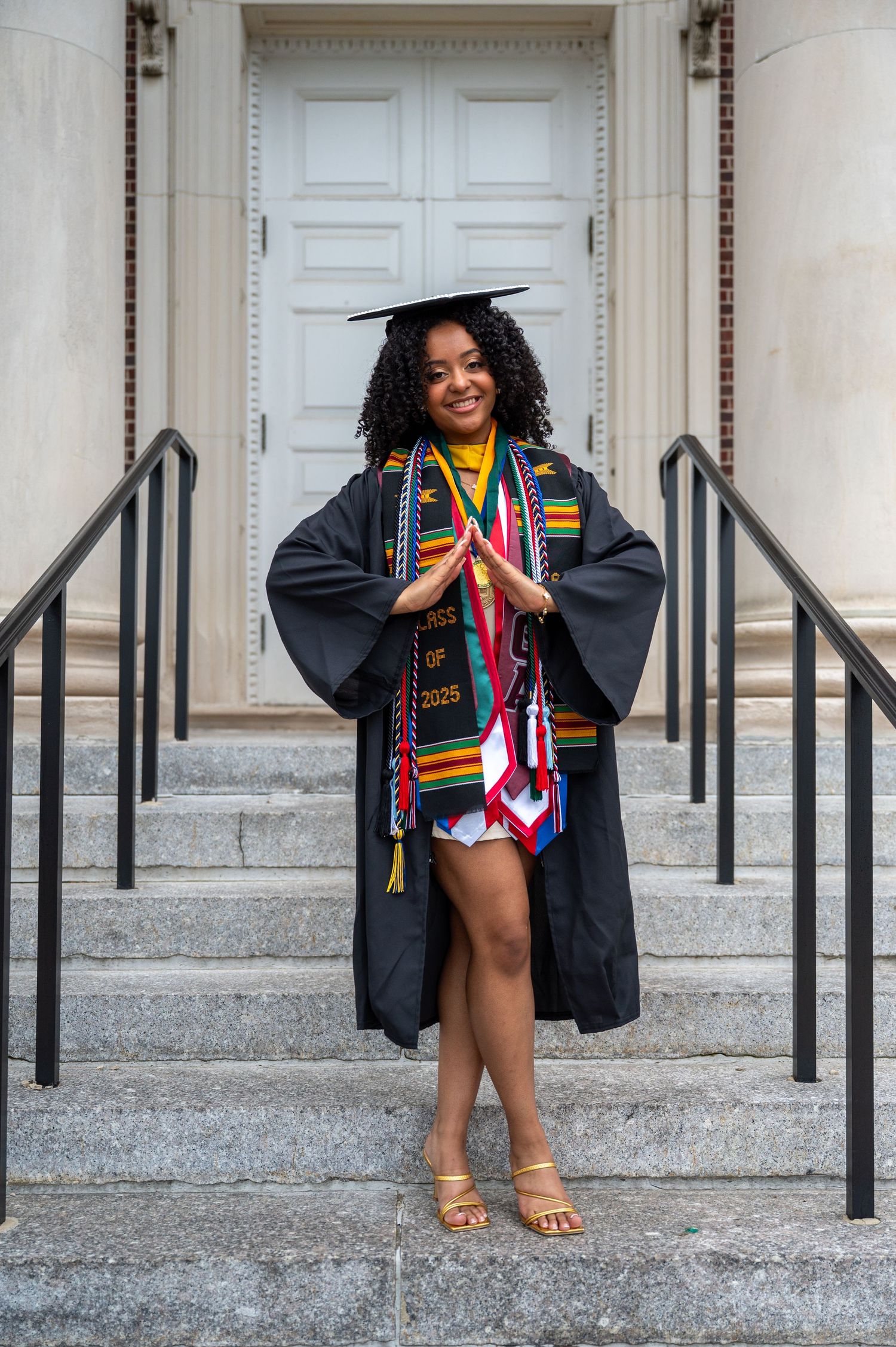 A graduation photoshoot on white stone steps featuring academic regalia and colorful stoles.