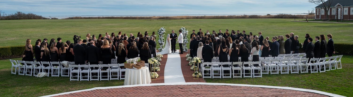 A panoramic view of an outdoor wedding ceremony setup with white chairs arranged on green grass under a blue sky.