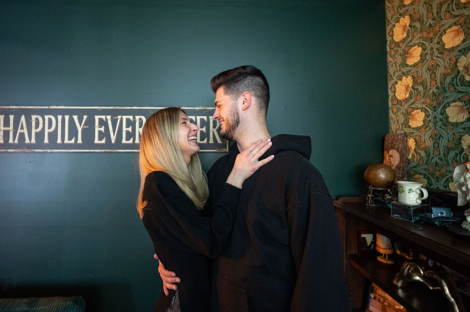 A couple shares an intimate moment in front of a wall with 'Happily Ever' text.