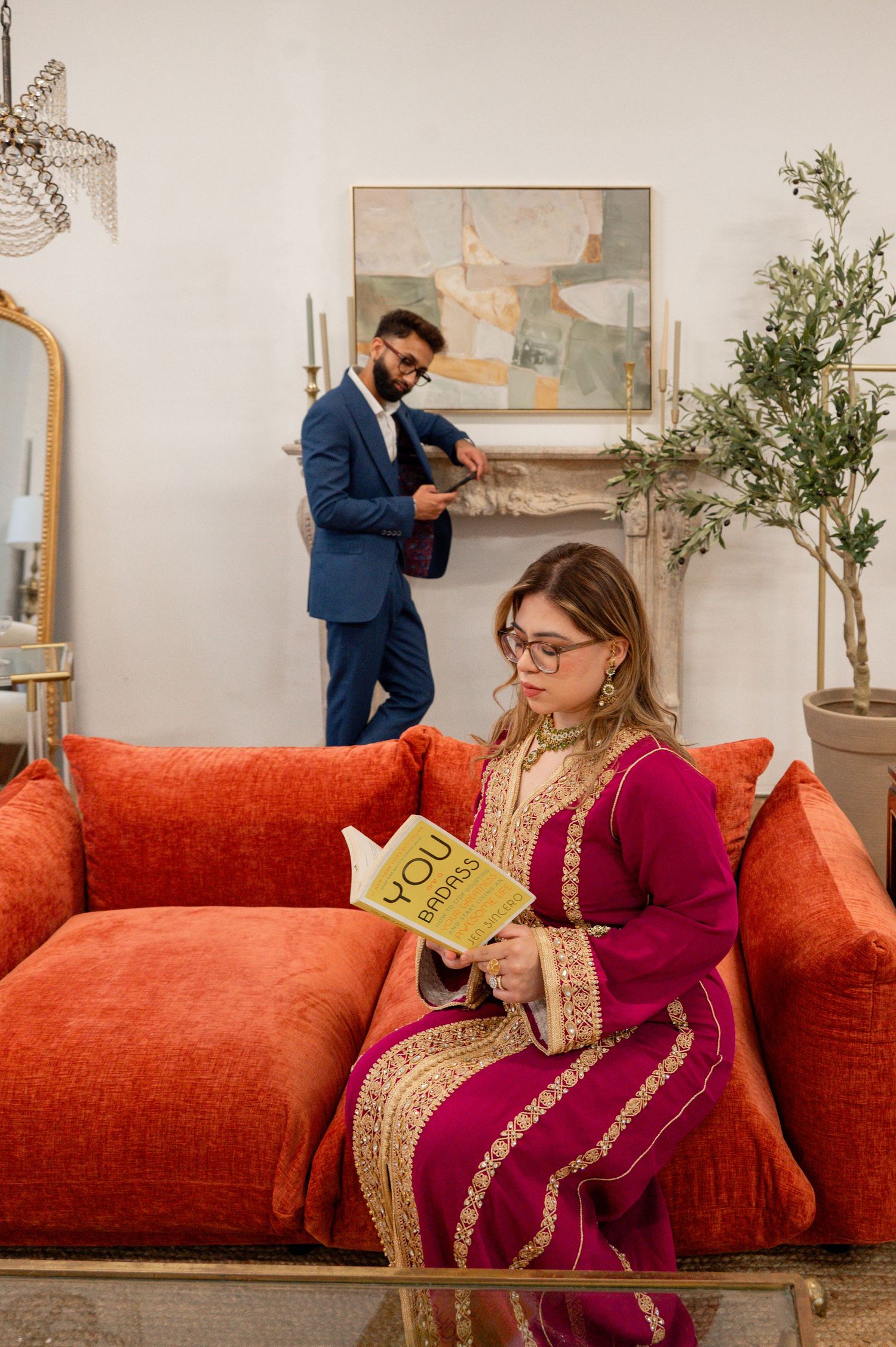 A couple in formal attire relaxes in a stylish living room with an orange velvet couch and decorative artwork.