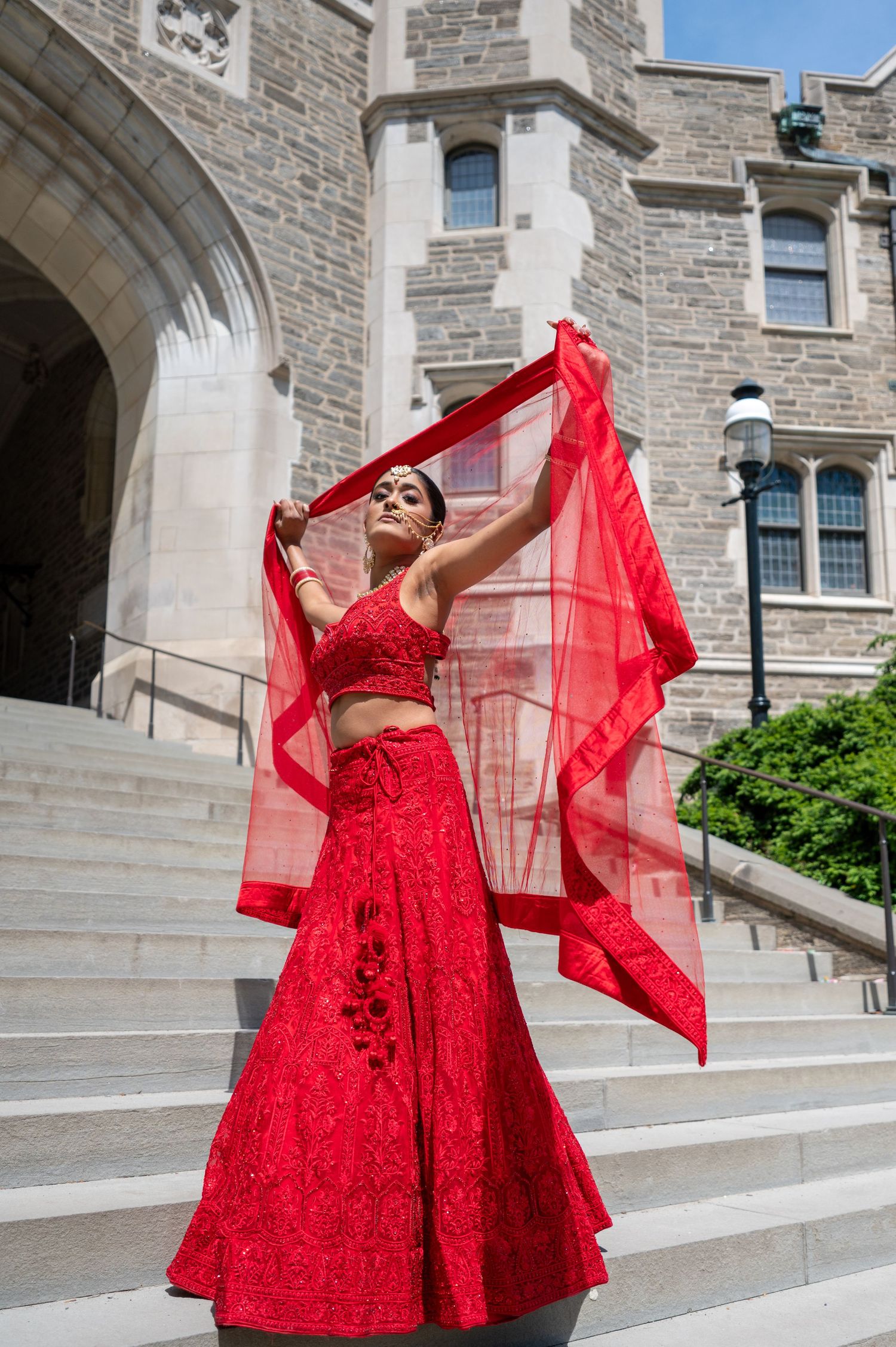 A stunning red lace gown with flowing sheer veil photographed on stone steps of a historic building.