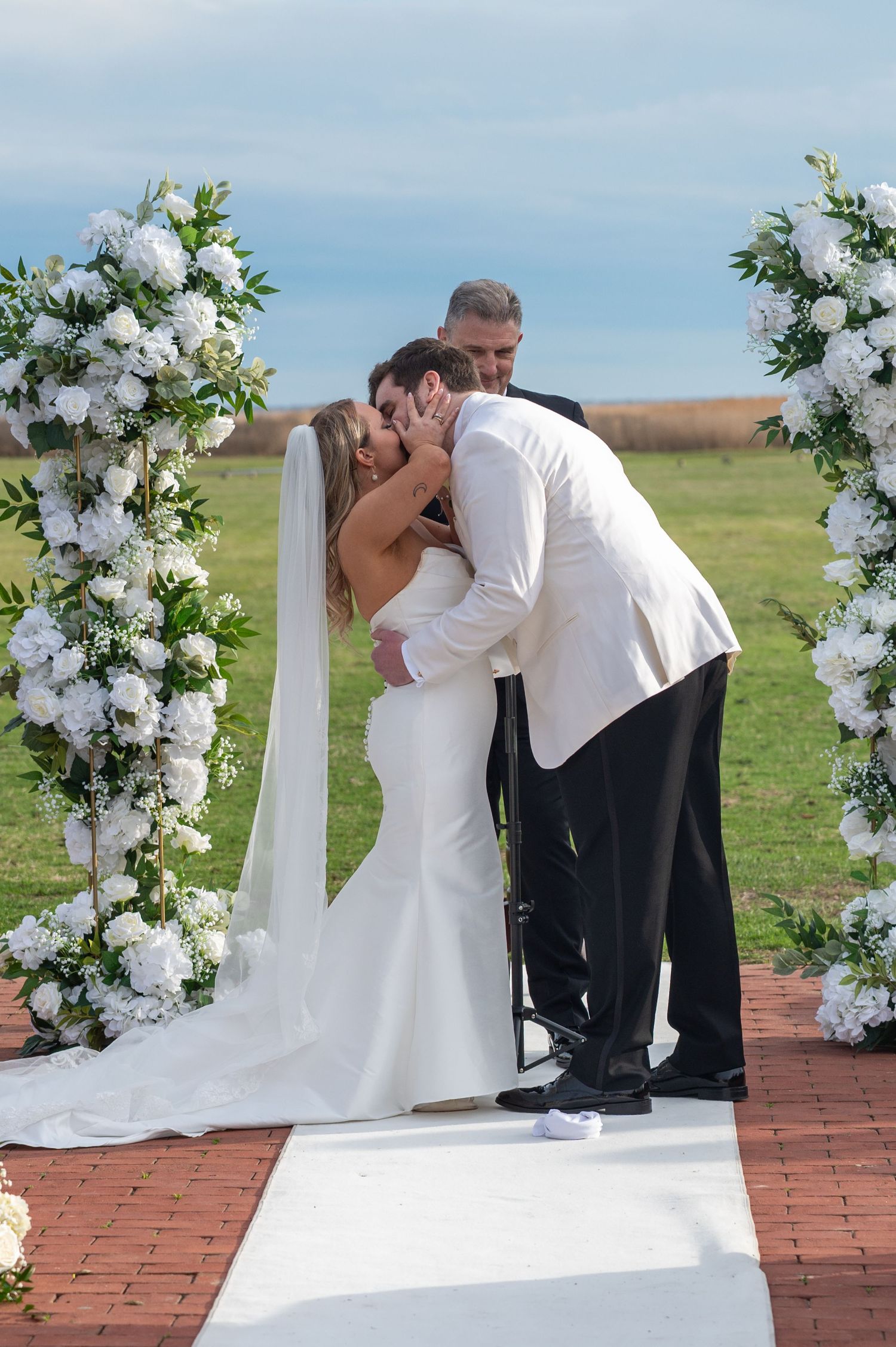 A wedding ceremony sequence showing a couple sharing their first kiss under a white floral arch on a brick patio.
