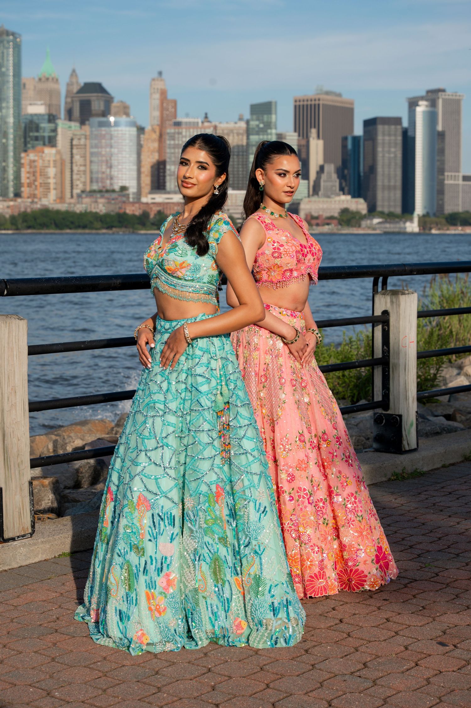 Models in turquoise and coral lehengas pose along waterfront with Manhattan skyline backdrop.