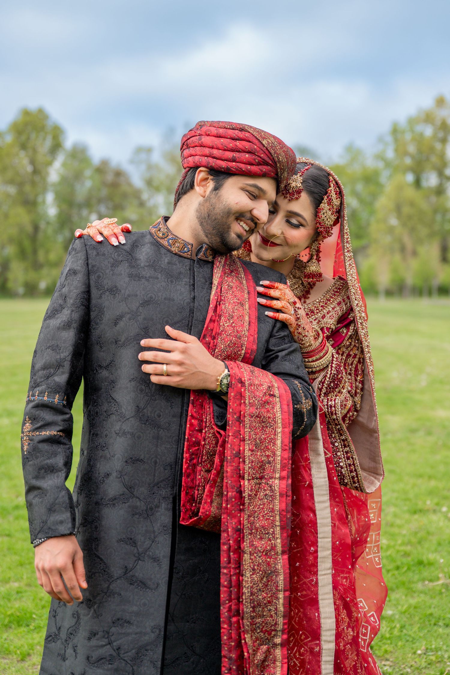 South Asian couple in traditional wedding attire embrace outdoors, groom in black sherwani and red turban, bride in red lehenga.