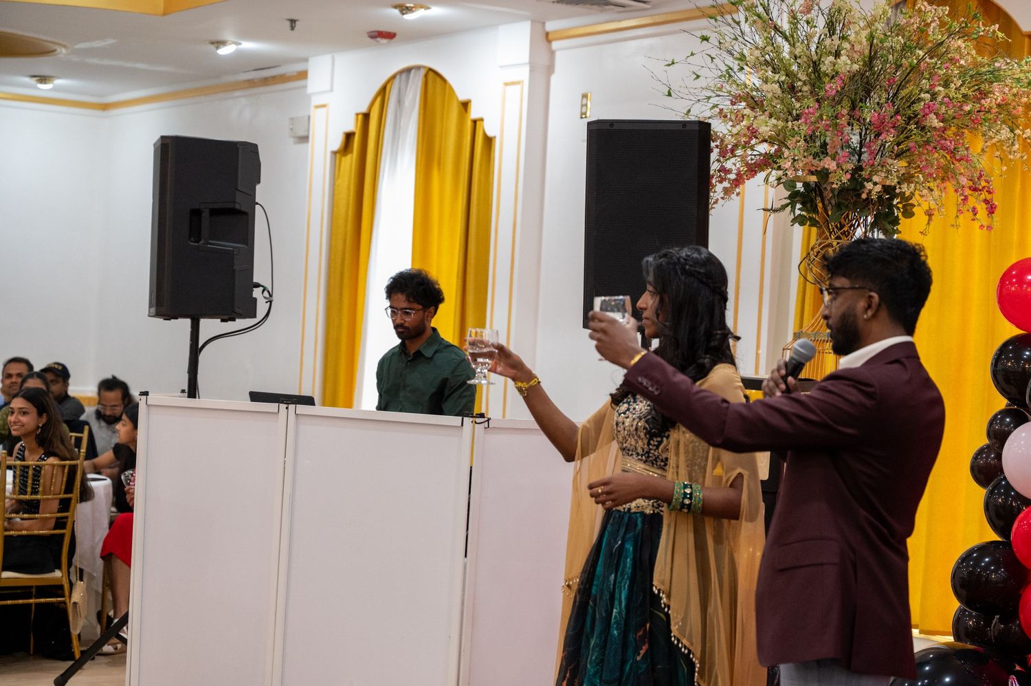 Performers on stage with speakers and floral decorations at a festive celebration.