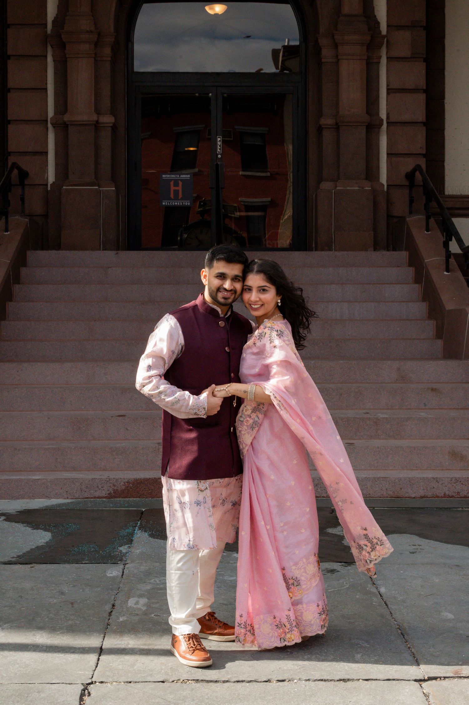 A couple in traditional South Asian attire poses lovingly on stone steps outside a building.