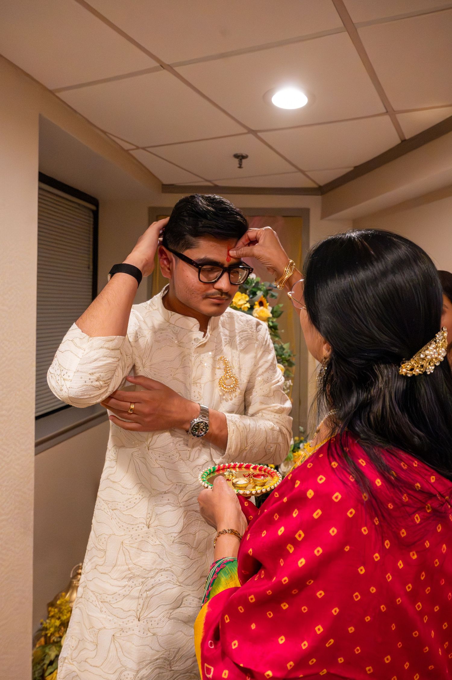 Traditional Indian ceremony shows a blessing ritual with people in white and red attire under indoor lighting.