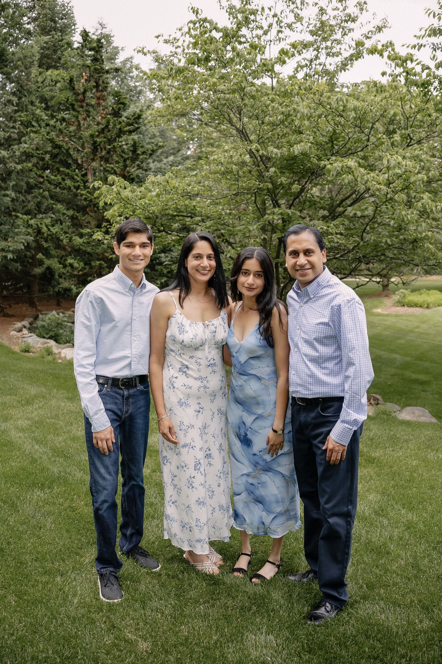 Family group posing together on green lawn with trees in background during summer photoshoot.