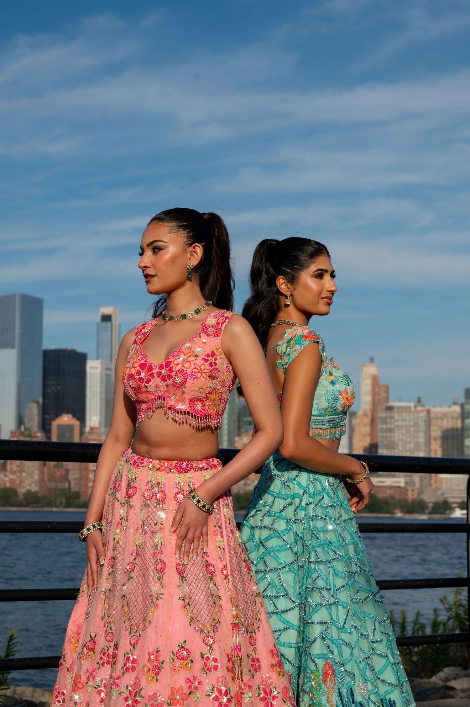 Two women in elegant Indian lehengas pose gracefully by a waterfront with city buildings in background.
