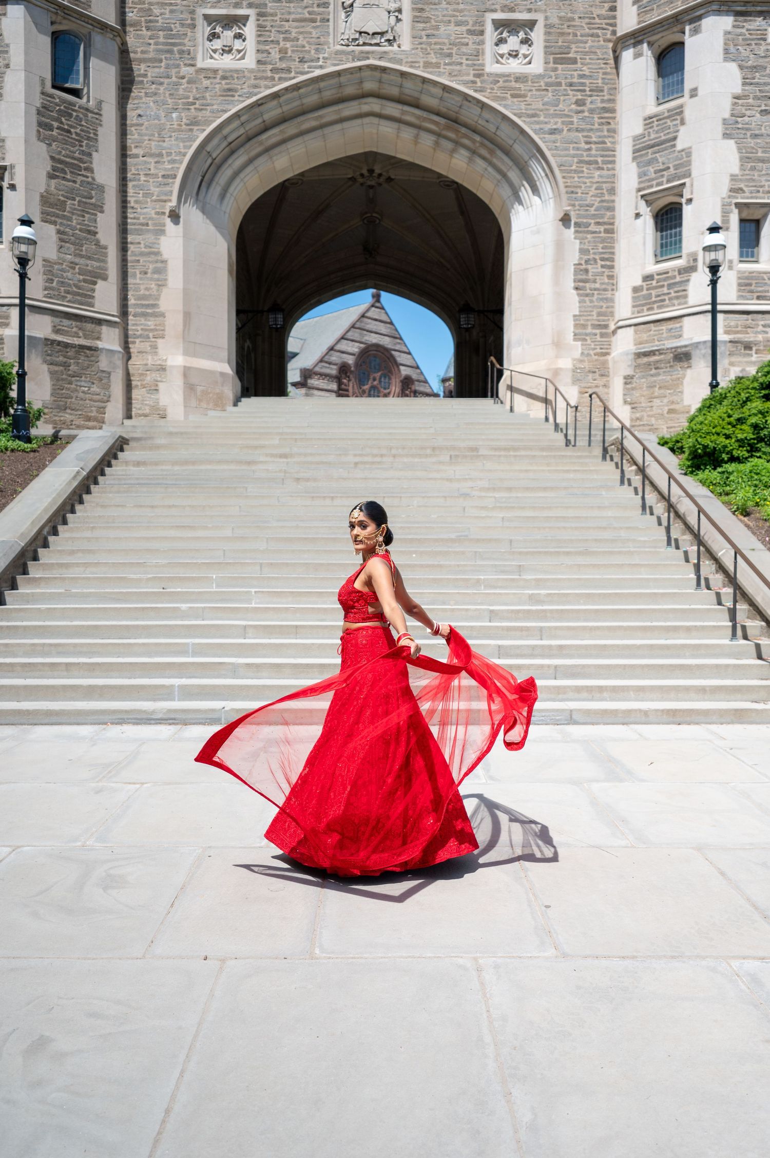 A dramatic red ball gown twirls on white stone steps in front of a Gothic architecture building.