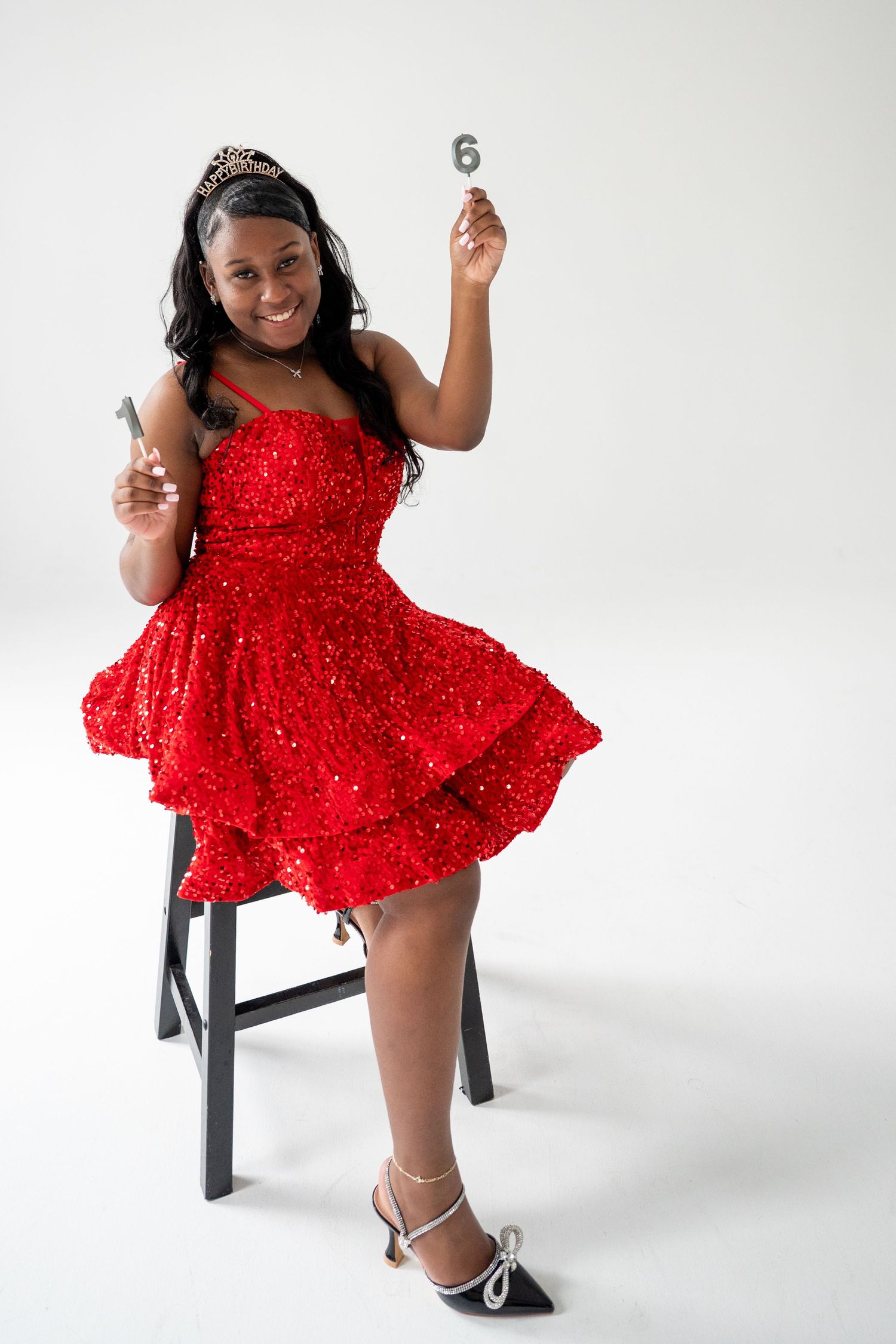Figure in sparkly red dress poses elegantly on black stool against white backdrop.