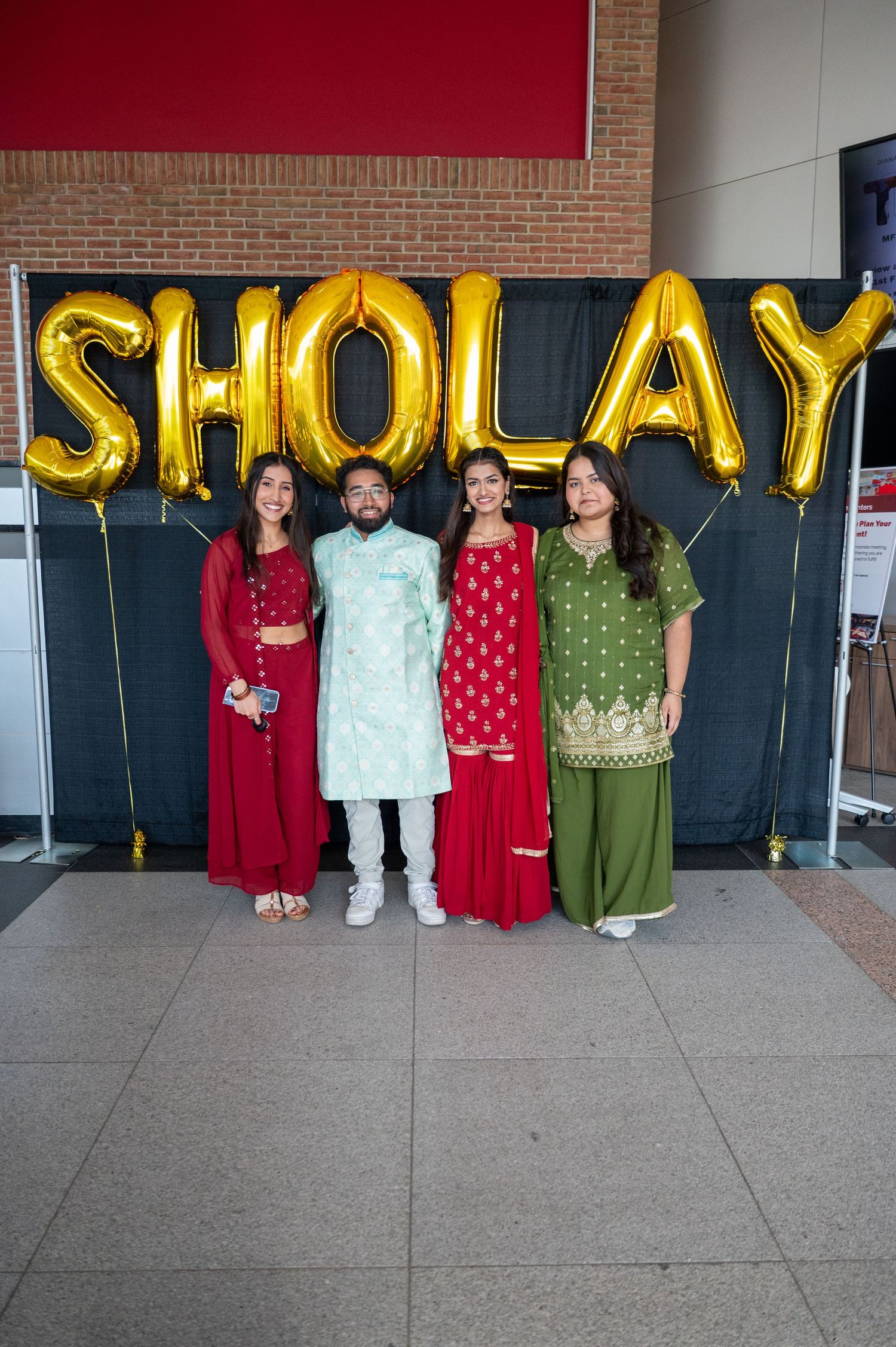 People in traditional cultural attire stand beneath gold letter balloons at an event.