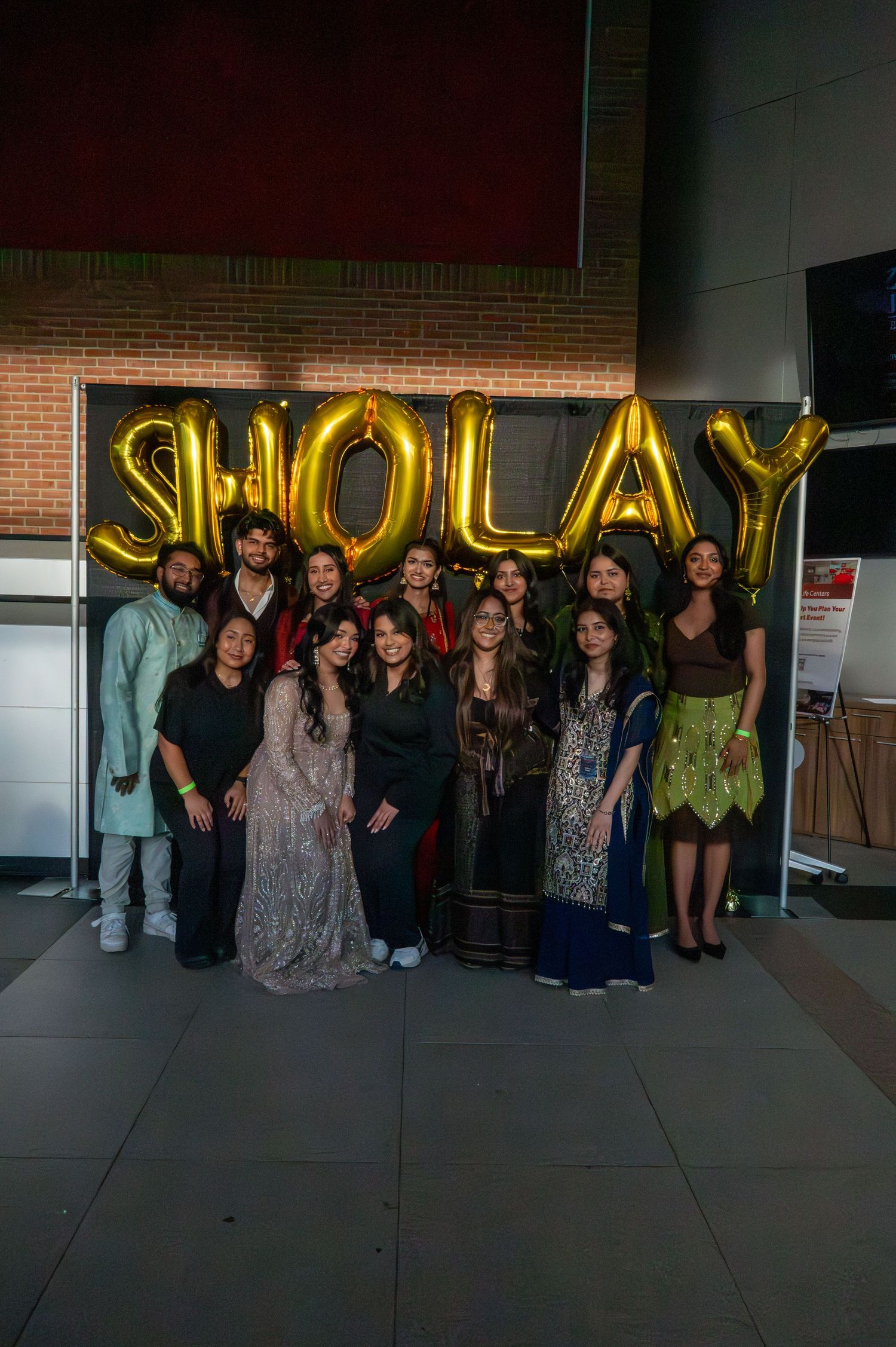 Group photo in front of golden balloon letters spelling SLAY with people in party attire.