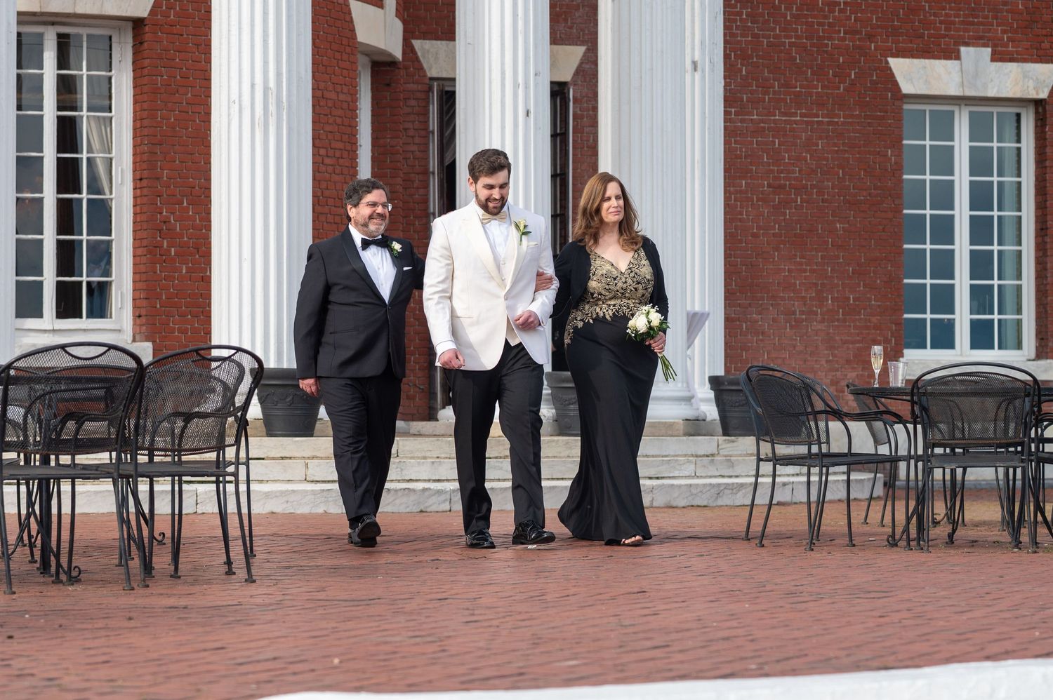 People in formal attire walking on a brick patio outside a stately building with white columns and windows.