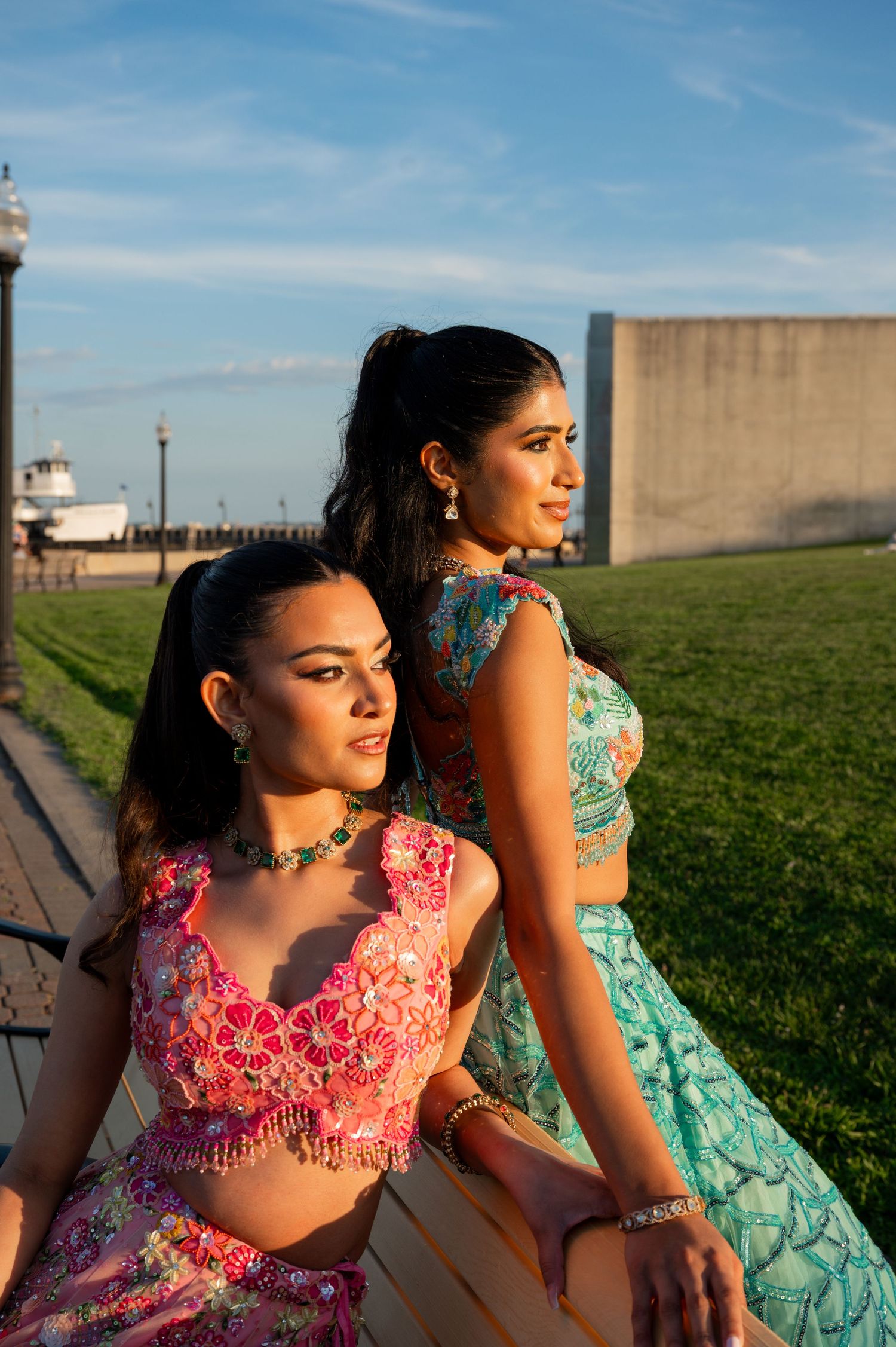 Two women in traditional Indian attire sit on bench during sunset photoshoot.