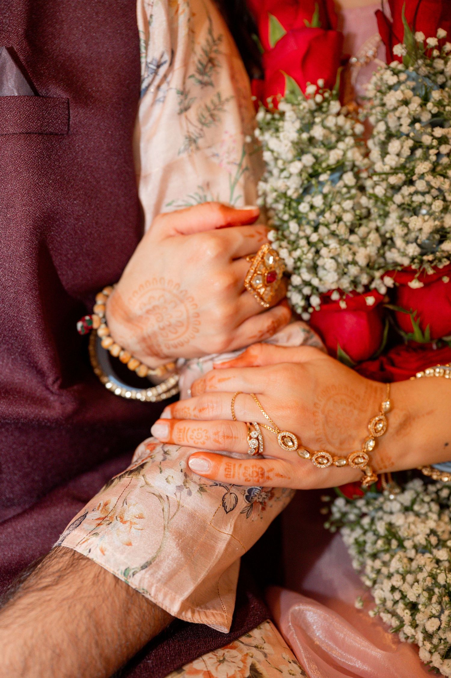 Close-up of hands with henna designs and gold jewelry holding delicate white baby's breath flowers against red fabric.