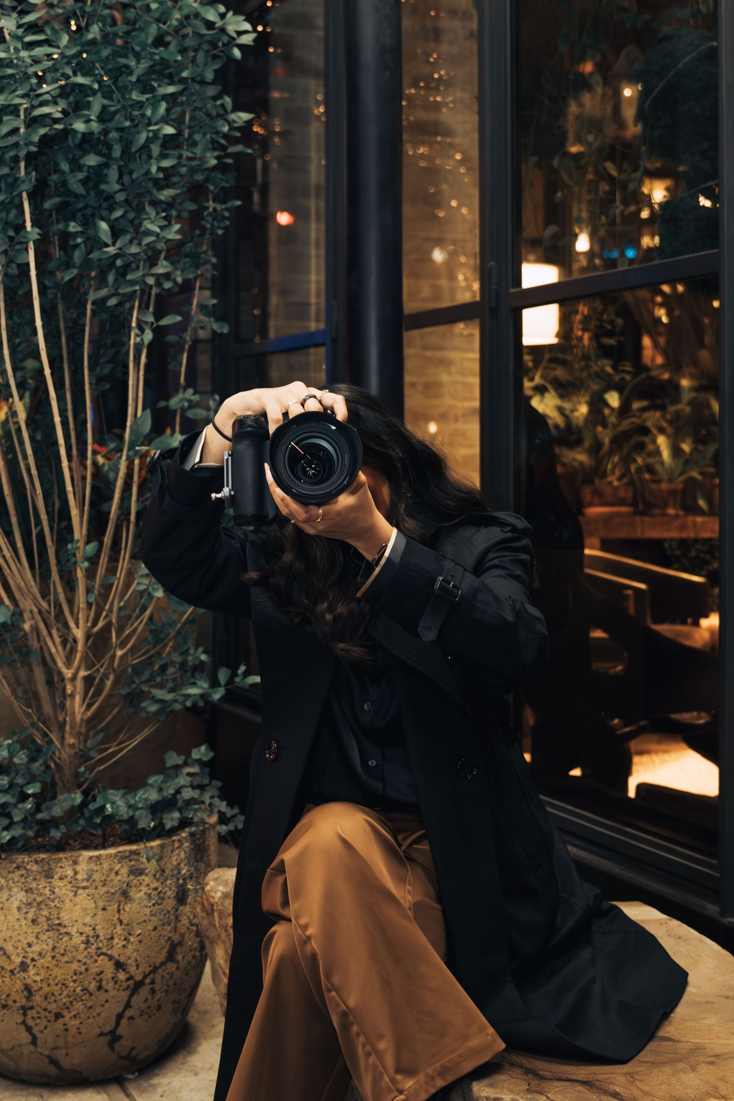 A photographer in a black coat takes photos while sitting next to a potted plant at dusk.