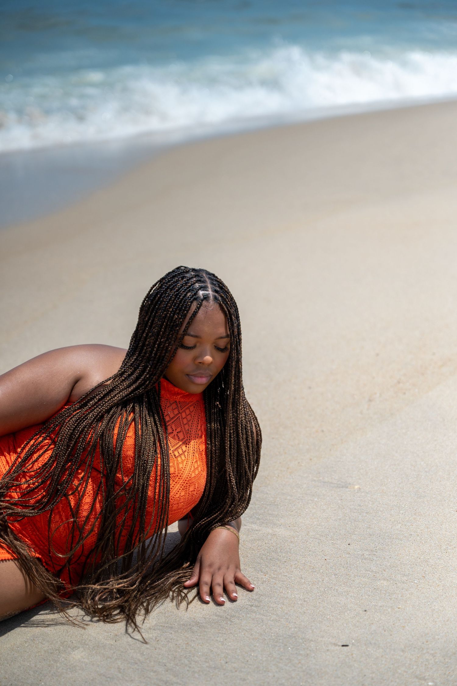 Model in orange dress poses dramatically on sandy beach with long braids flowing.