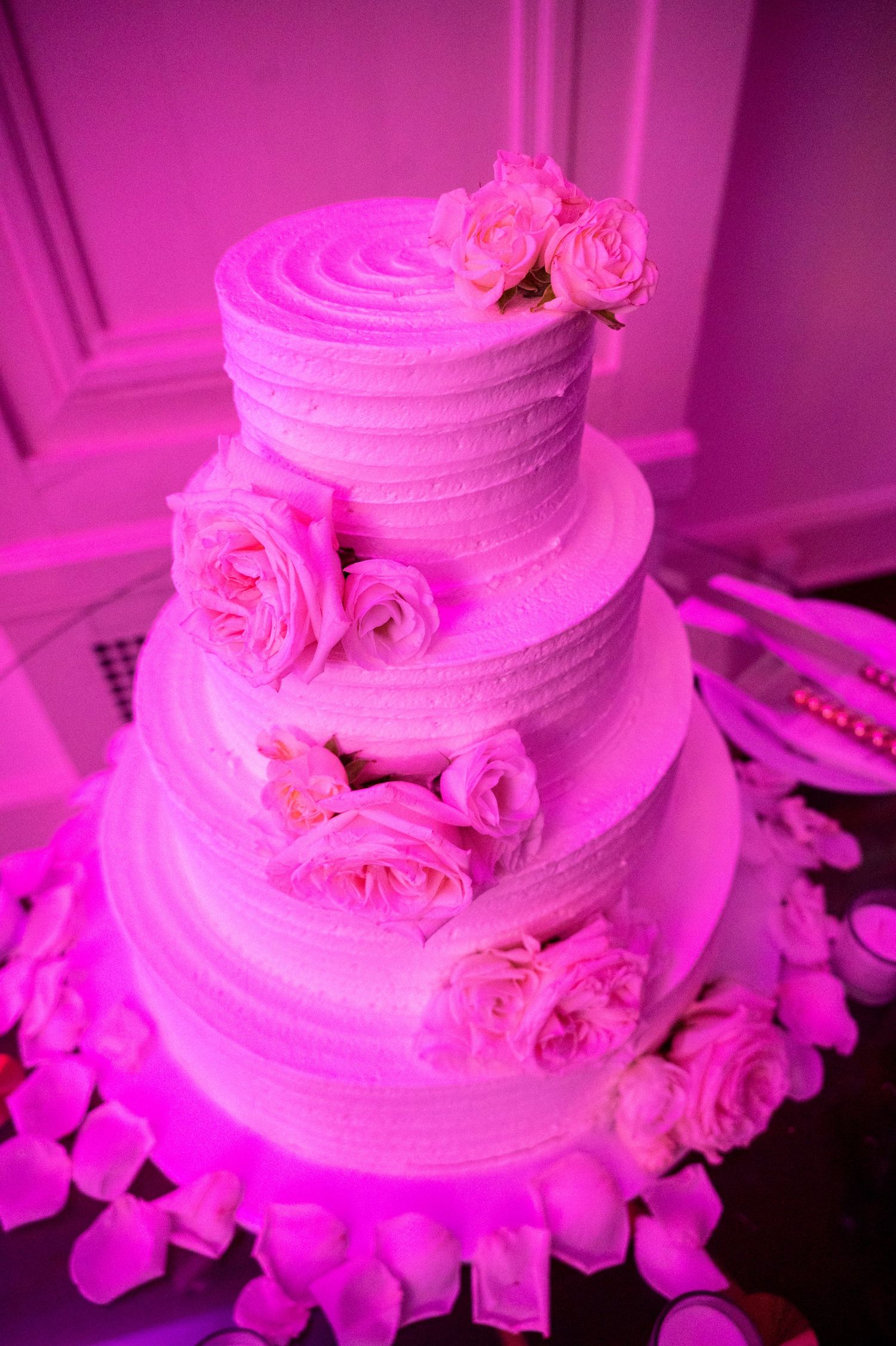 A white tiered wedding cake with pink roses and purple lighting creates a romantic atmosphere at a reception.