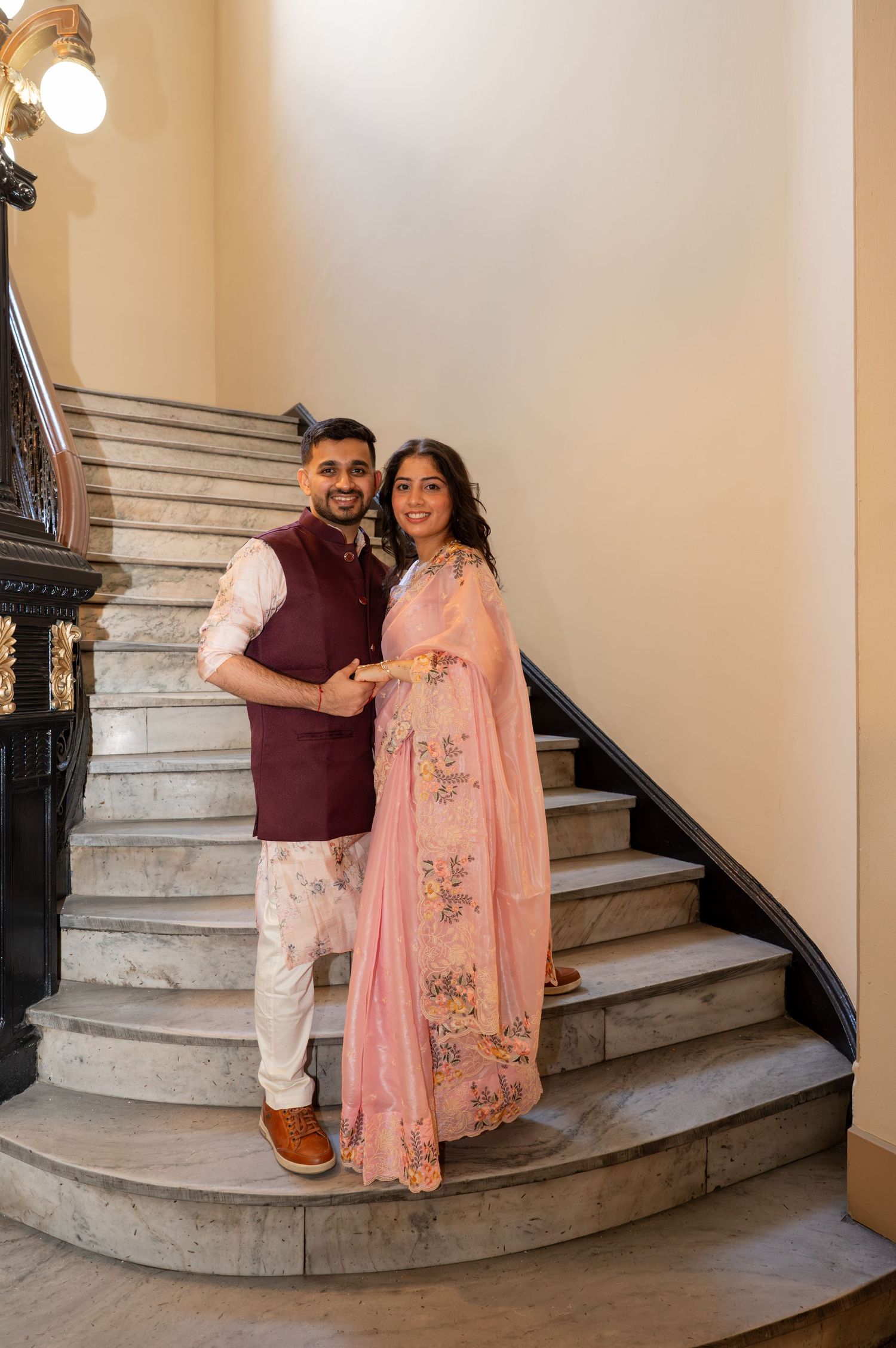 A couple poses on an elegant marble staircase in traditional South Asian attire.