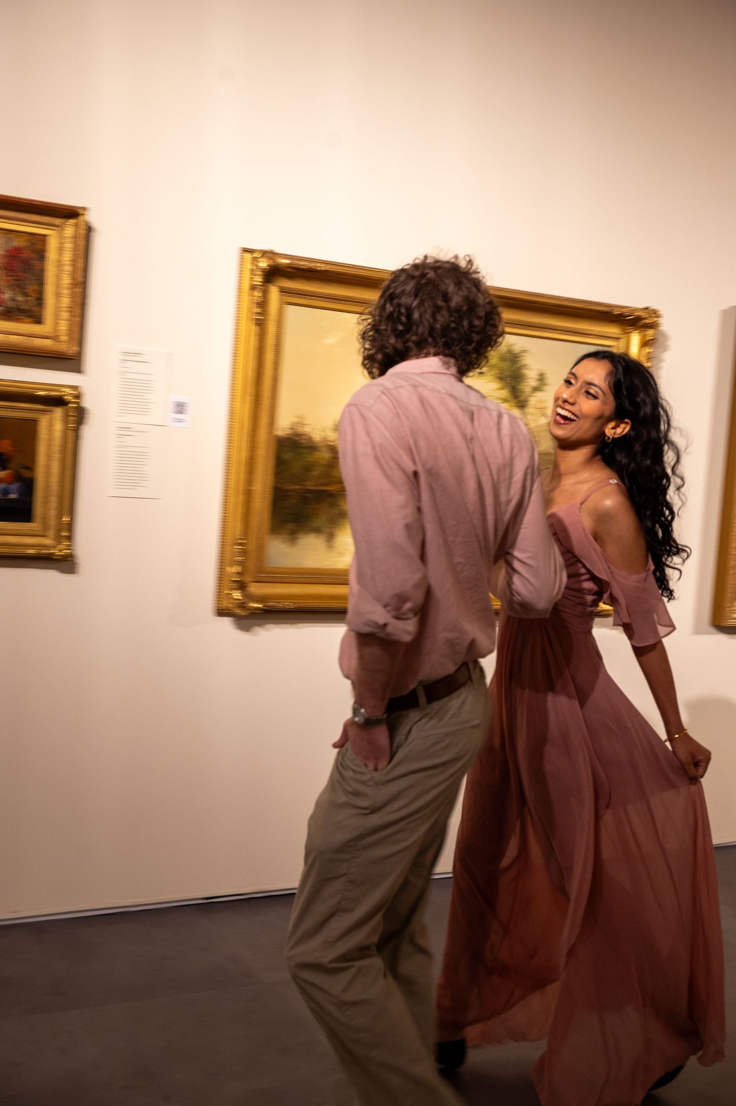 A couple dancing and laughing together in an art gallery with gold-framed paintings.