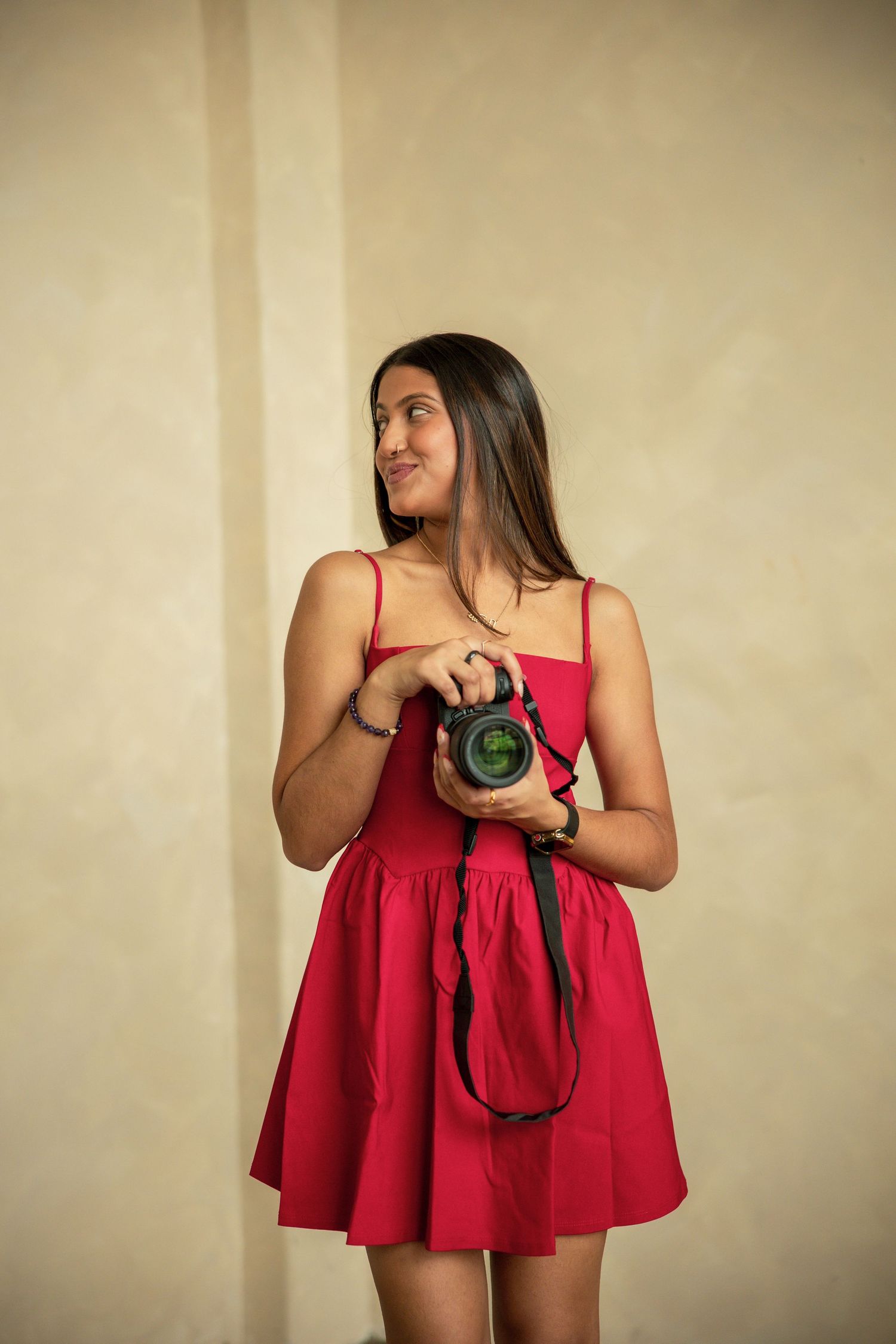 Woman in red dress poses playfully with camera against beige wall in artistic portrait series.