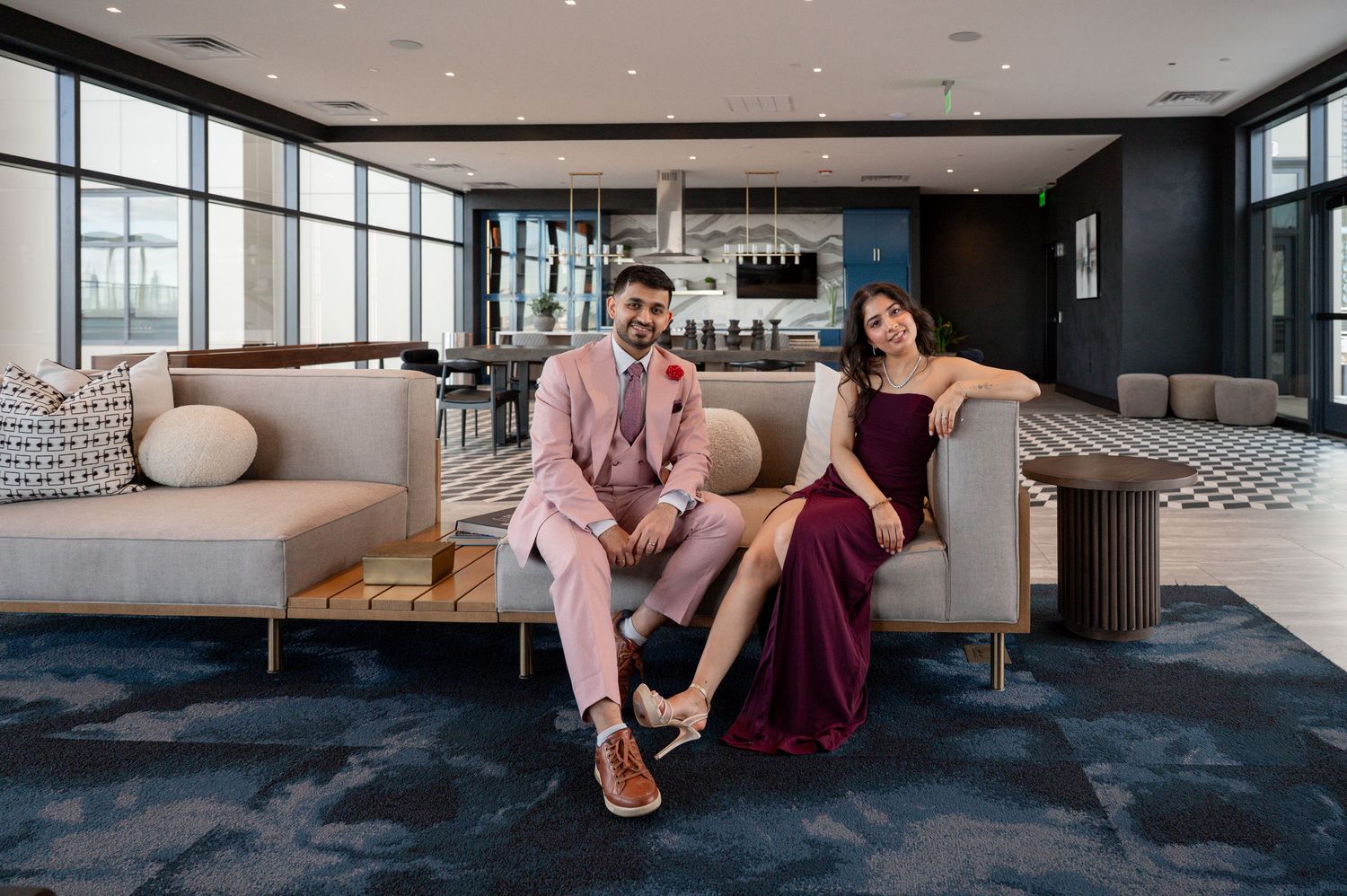 Two people in formal attire relax on a modern gray sofa in an upscale lounge area with dark flooring.