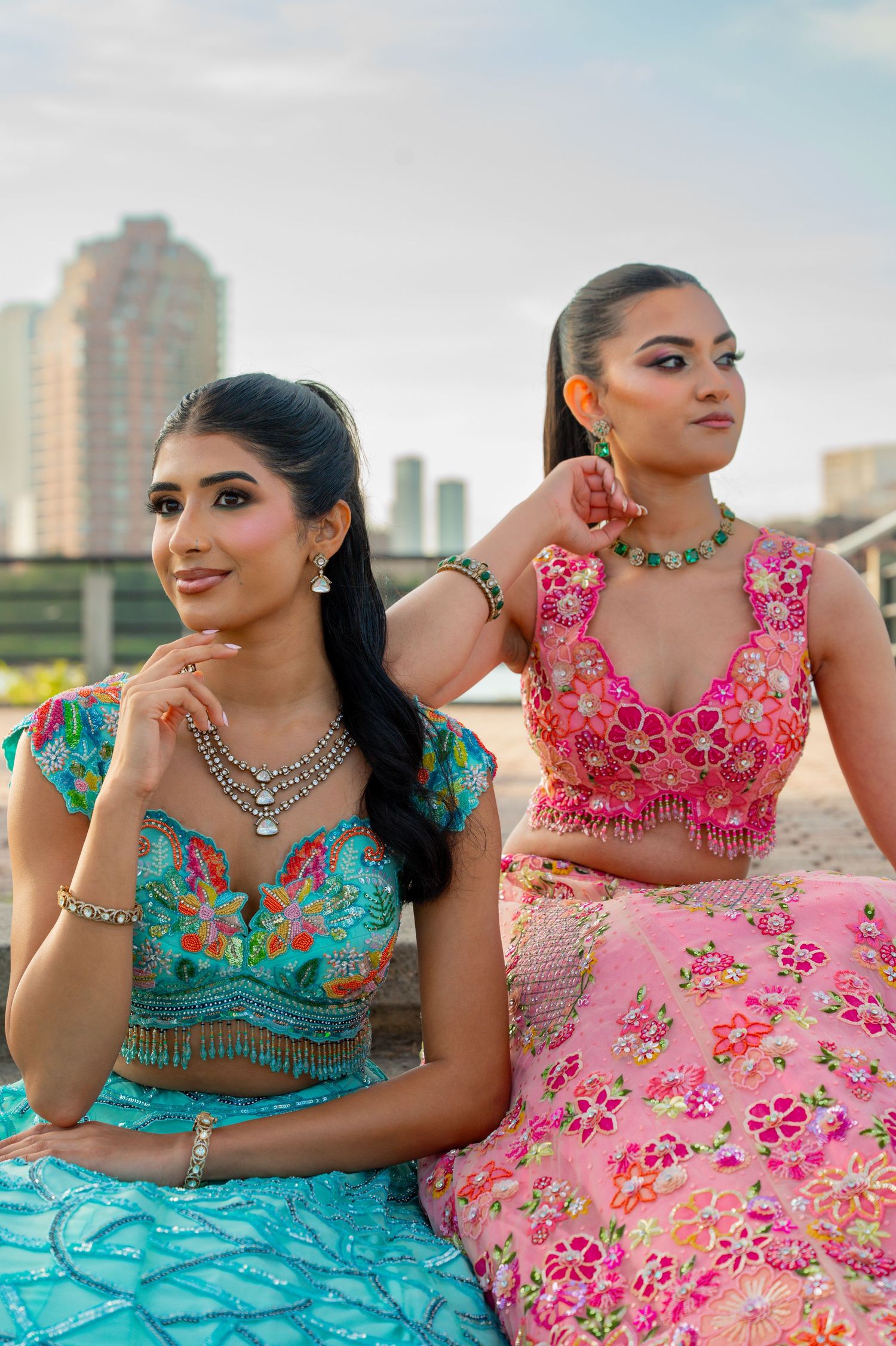 Two women in colorful floral lehengas posing by pool with city skyline backdrop.