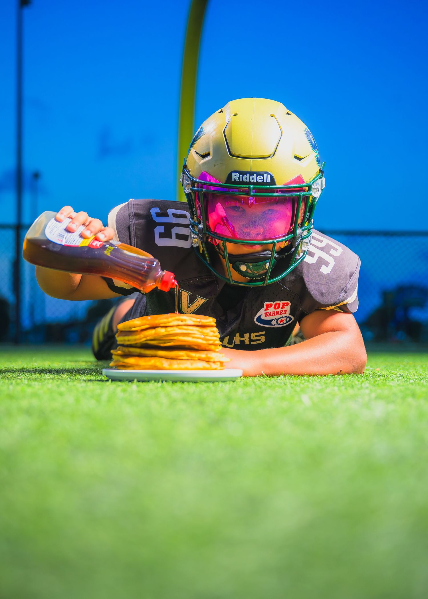 Football player wearing yellow helmet and visor pours syrup on stack of pancakes on artificial turf field.