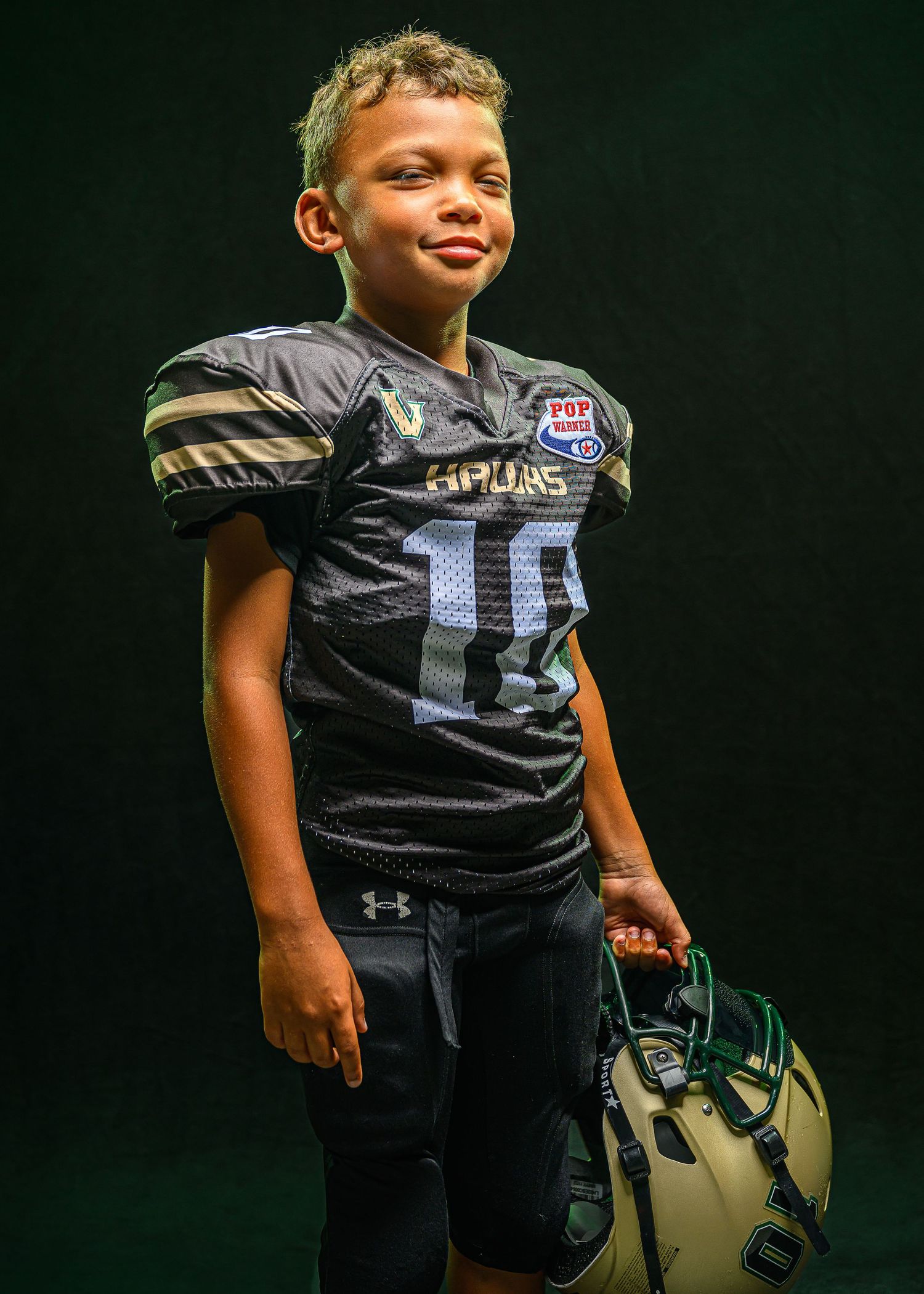 Young football player in black Under Armour jersey shows determination while posing with golden helmet.