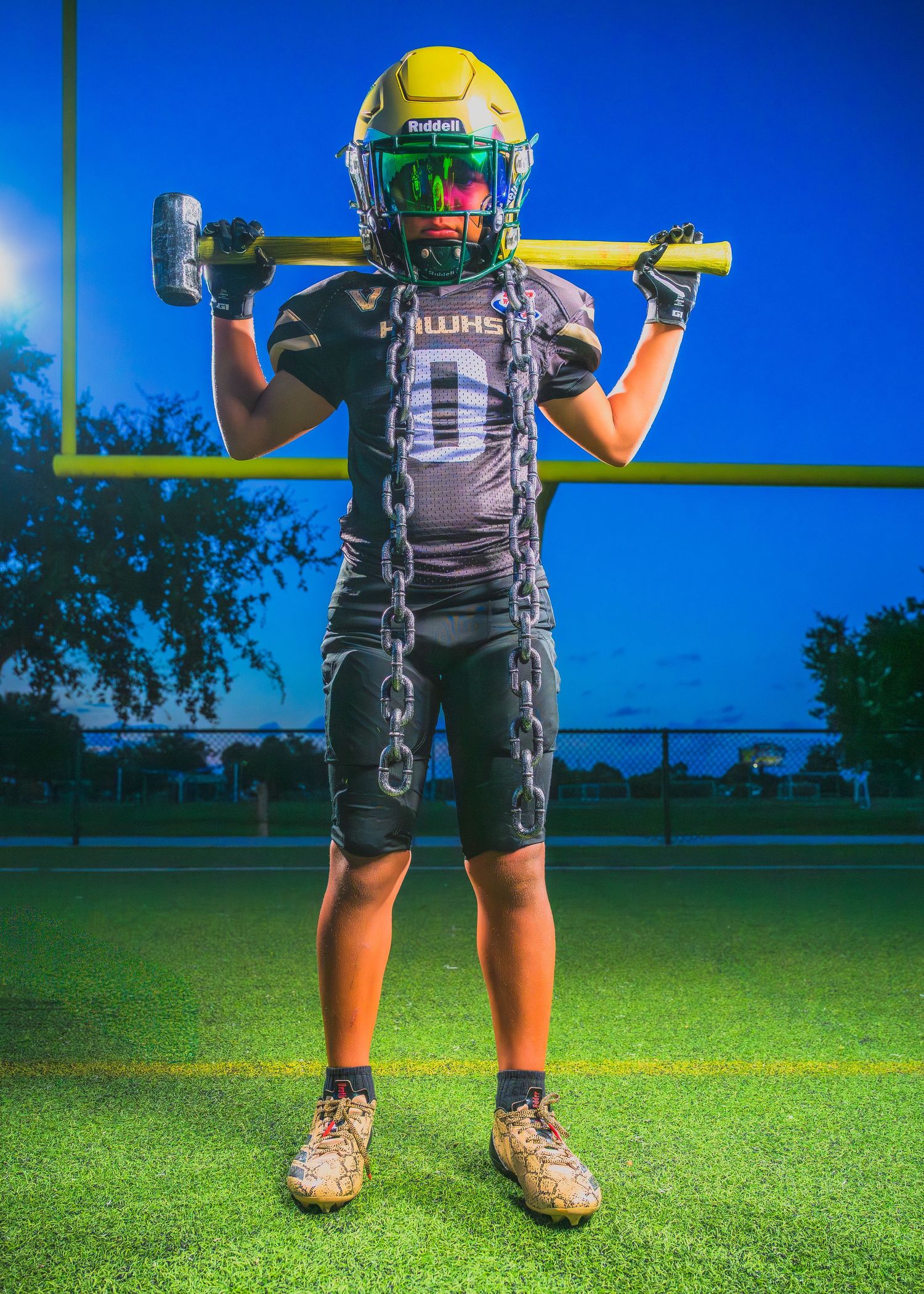 Football player wearing black uniform and yellow helmet poses with equipment on field at dusk against blue sky.