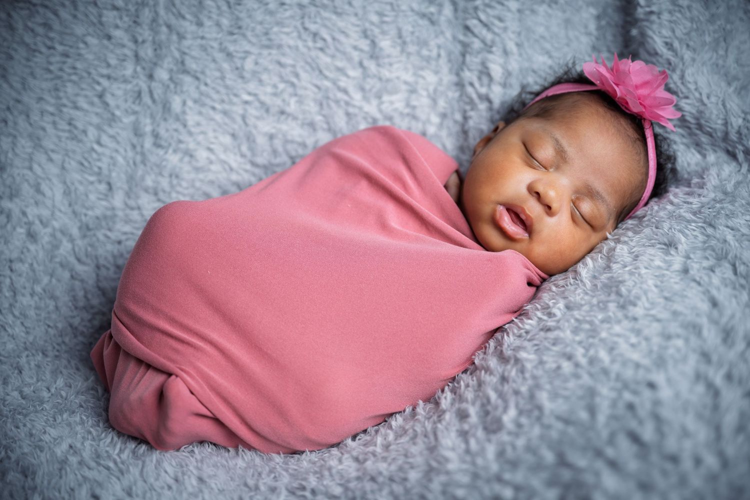 Sleeping newborn wrapped in pink swaddle on grey textured blanket.