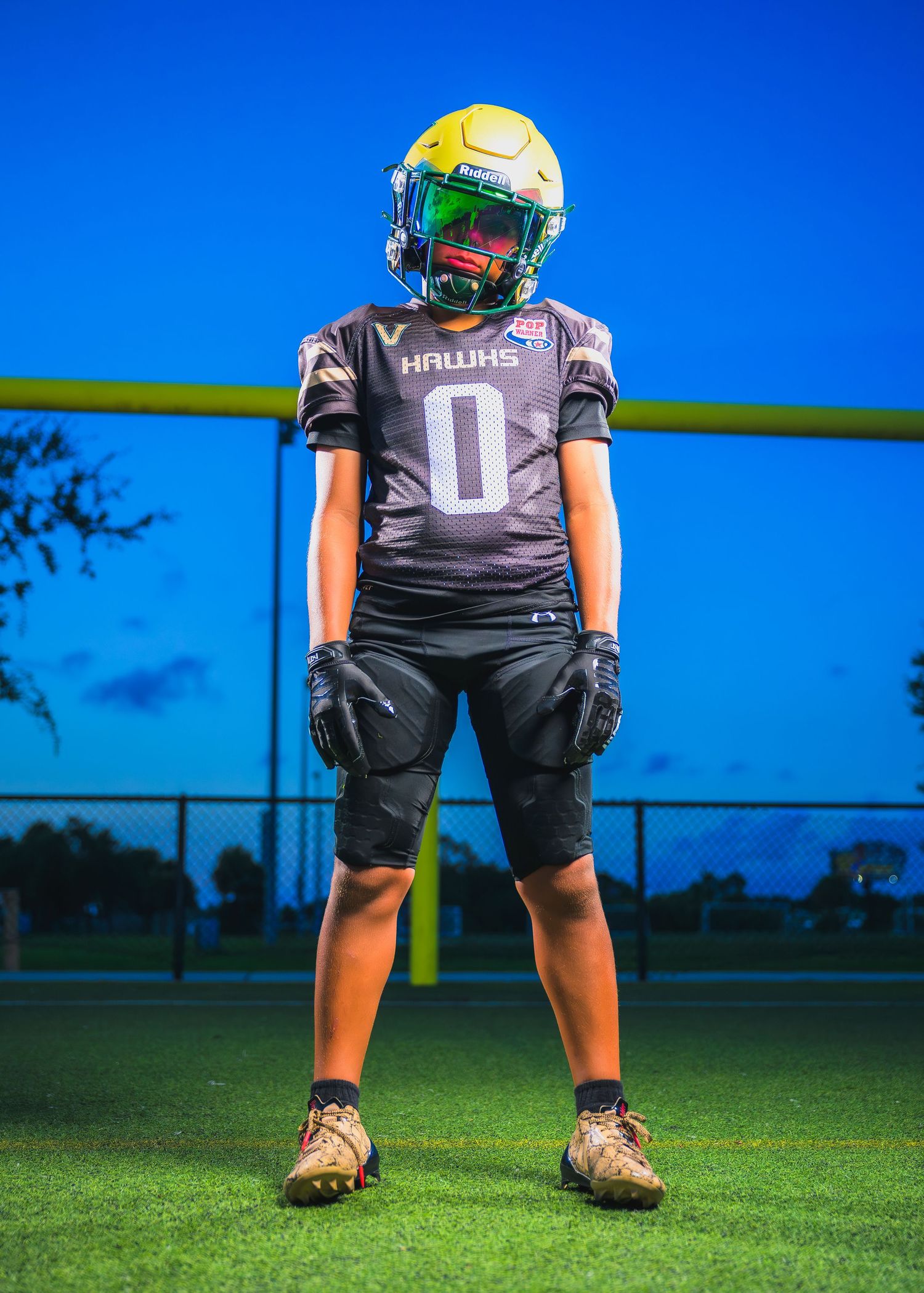 Youth football player wearing gray jersey number 0 and yellow helmet stands ready on artificial turf field at dusk.