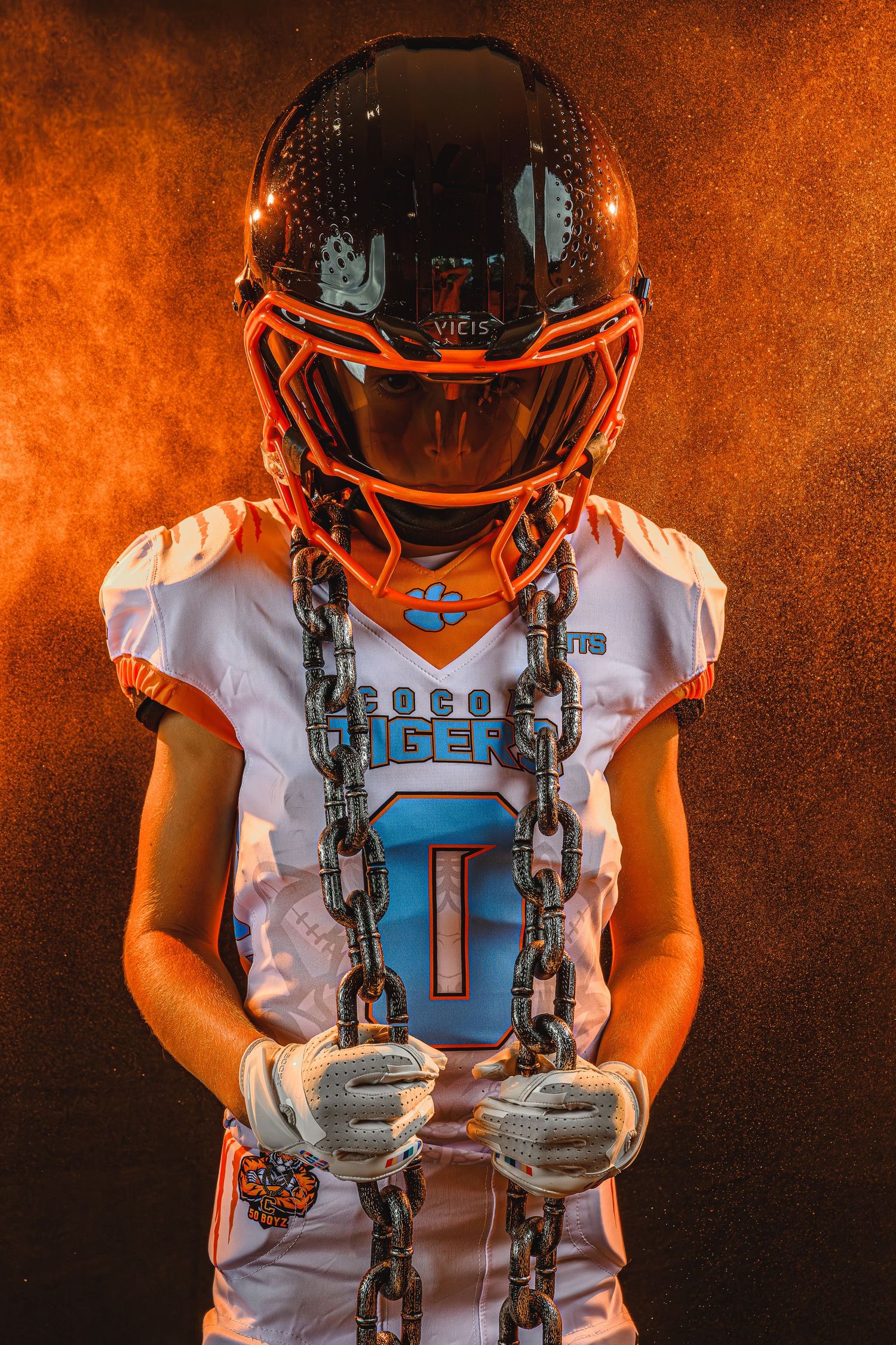 Football player wearing white jersey with orange helmet holds a gun-shaped prop against dark orange background.