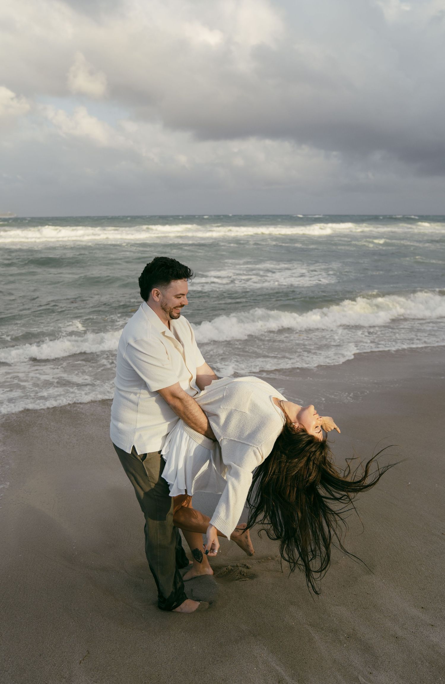 A person in white clothing dances with their long hair flowing on a stormy beach against dramatic waves and clouds.