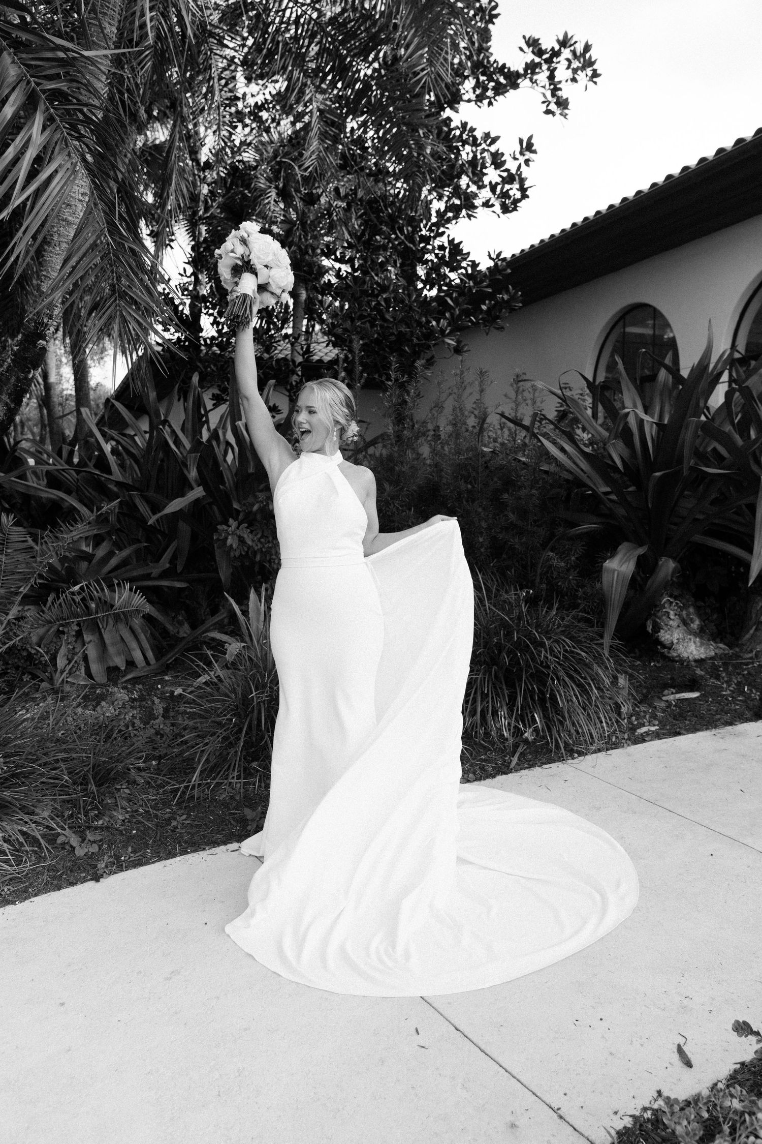 A graceful bride in a flowing white wedding dress twirls and spins across a light-colored stone patio in a garden setting.